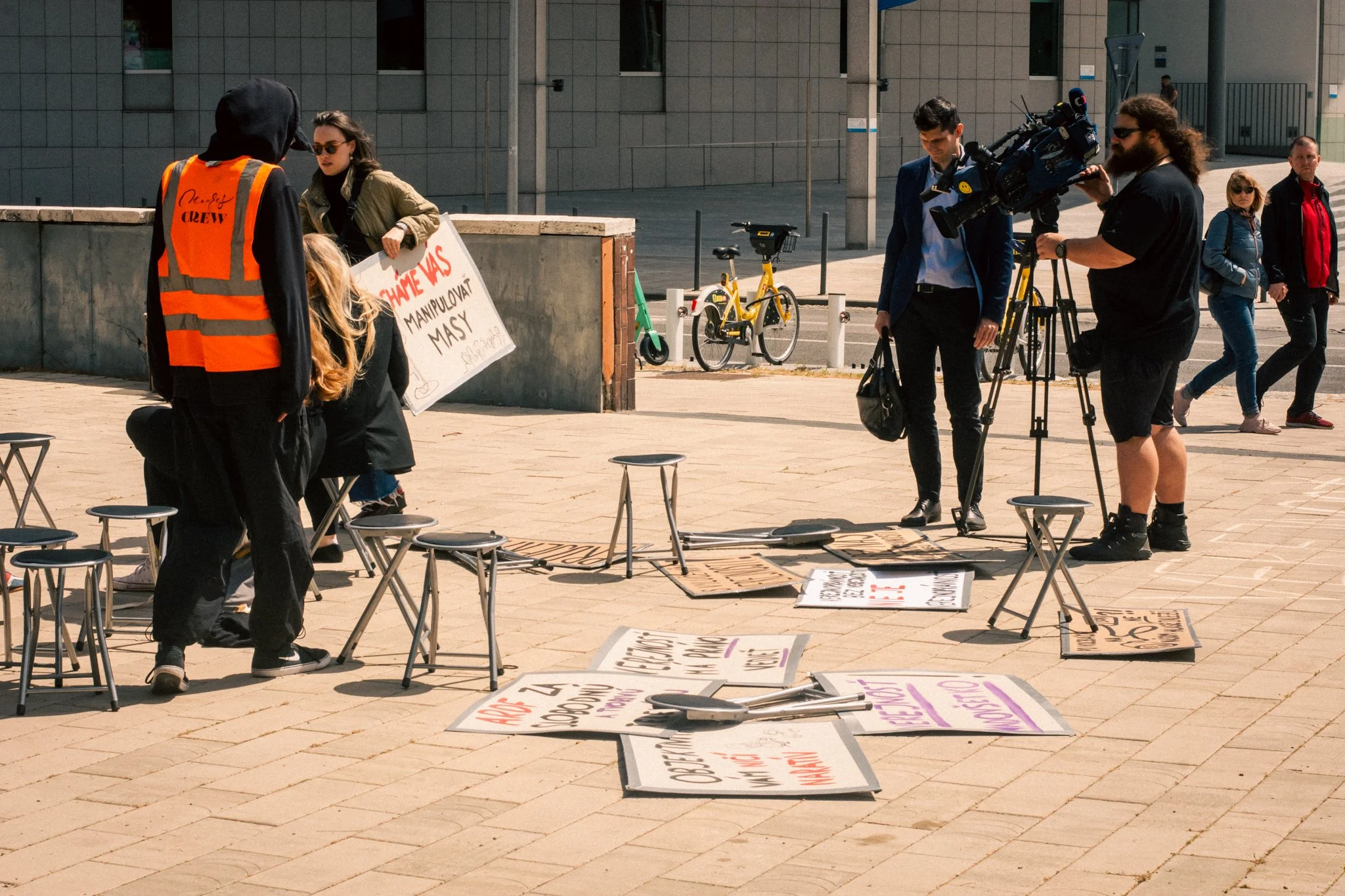 Protest on a sidewalk with signs and a television crew filming, several people standing and walking in the background.