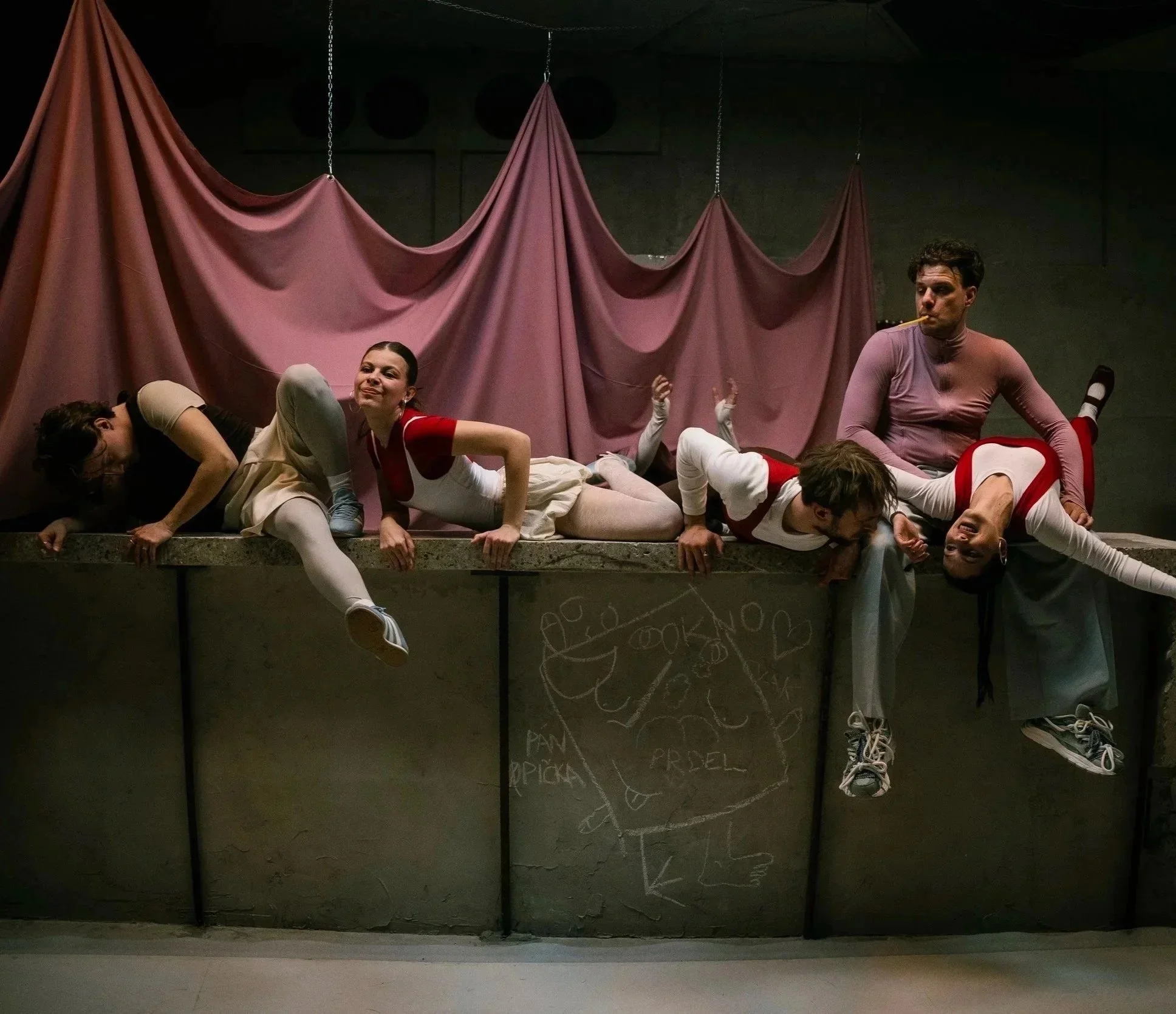 Actors in a theatrical performance lying upside down on a raised platform with a pink curtain backdrop, some with playful expressions and props.