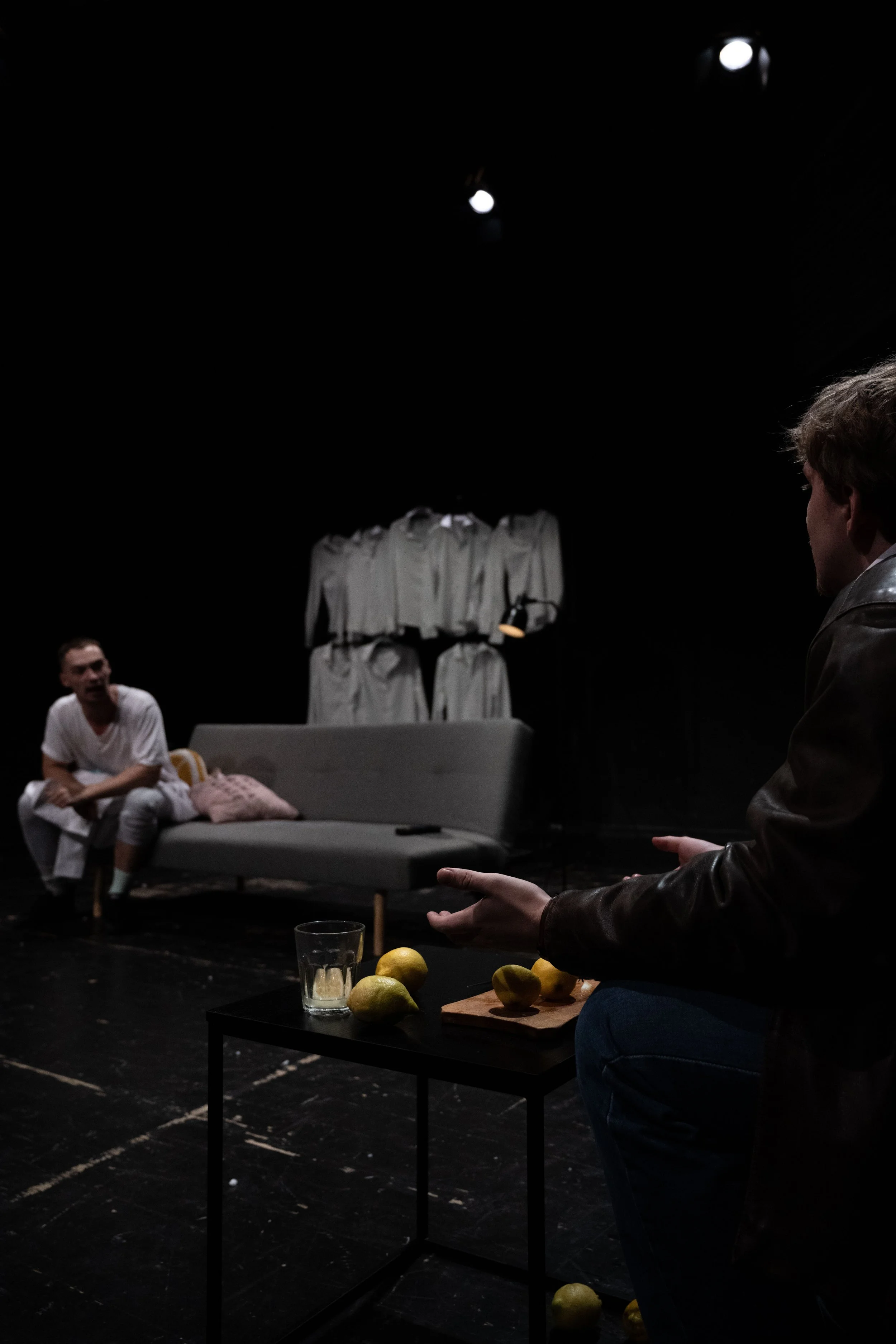 Two people are on a dark stage set, with one sitting on a couch and the other sitting nearby, gesturing. There are lemons on a small table, a glass of water, and a clothing rack with gray suits in the background.