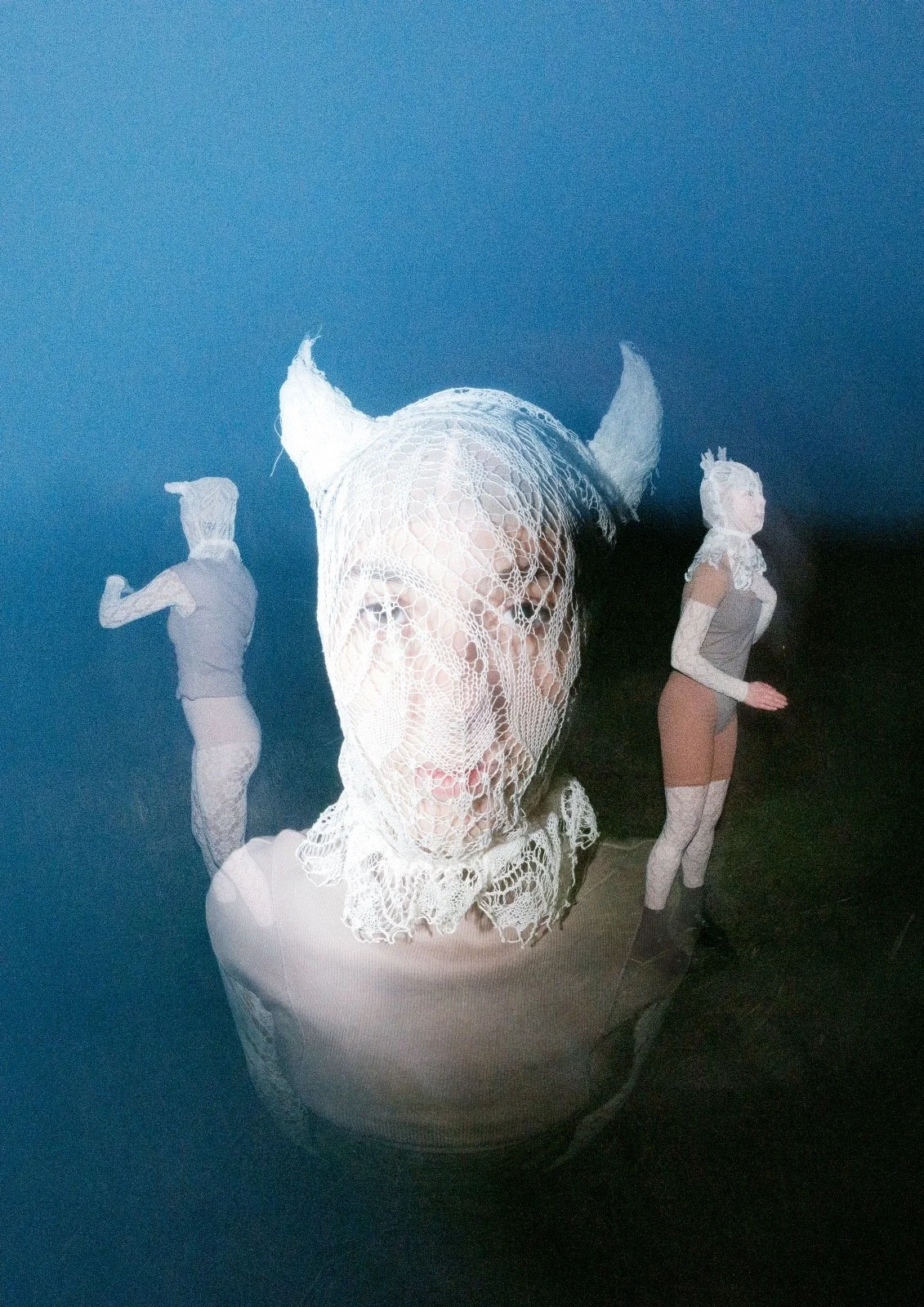 Person wearing a fishnet hood with horns, flanked by two women in stockings and bustiers, standing against a dark background.