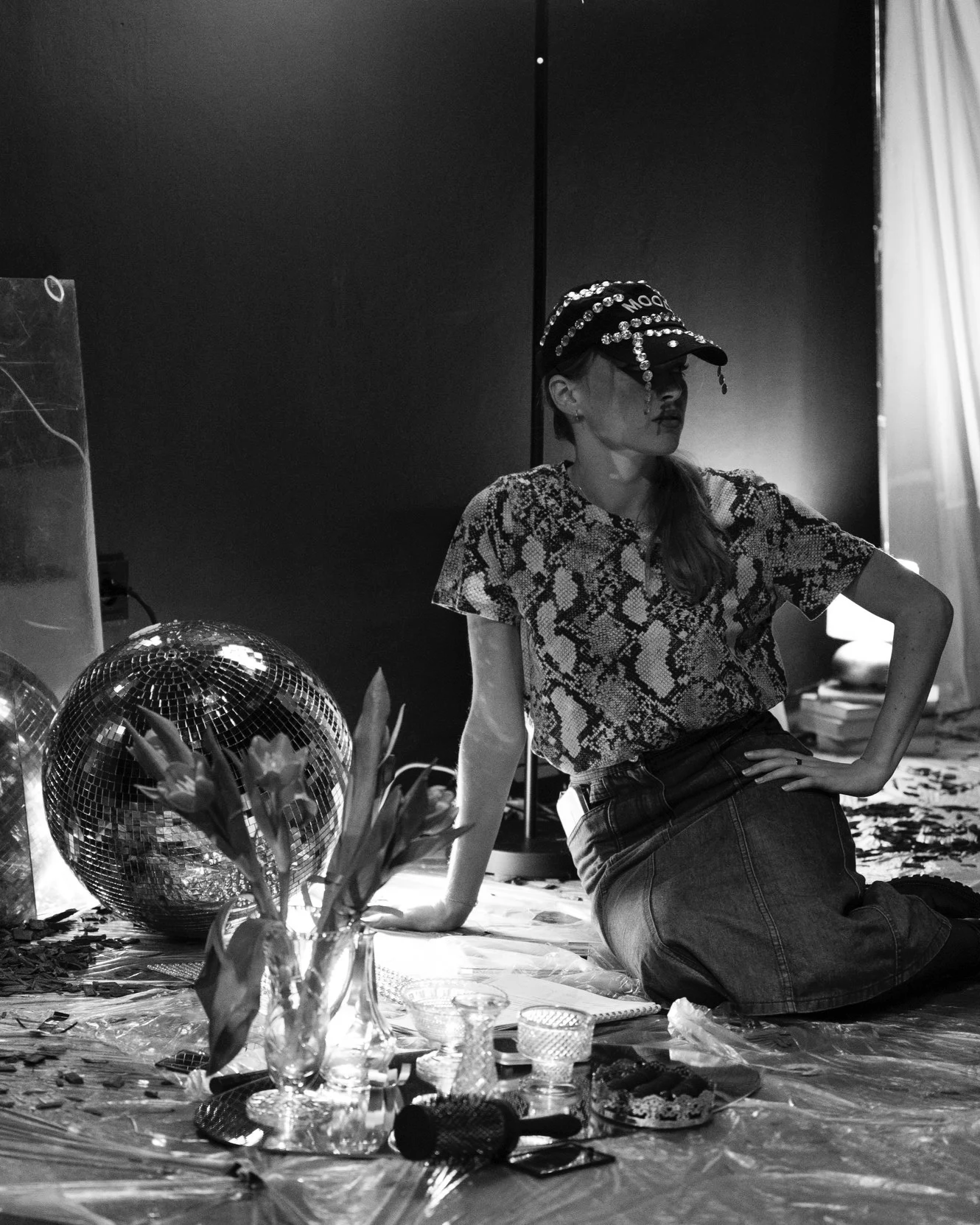 A woman sitting on the floor, wearing a patterned cap and snake print shirt, near a disco ball, flowers in a vase, and various items on the ground in a dimly lit room.