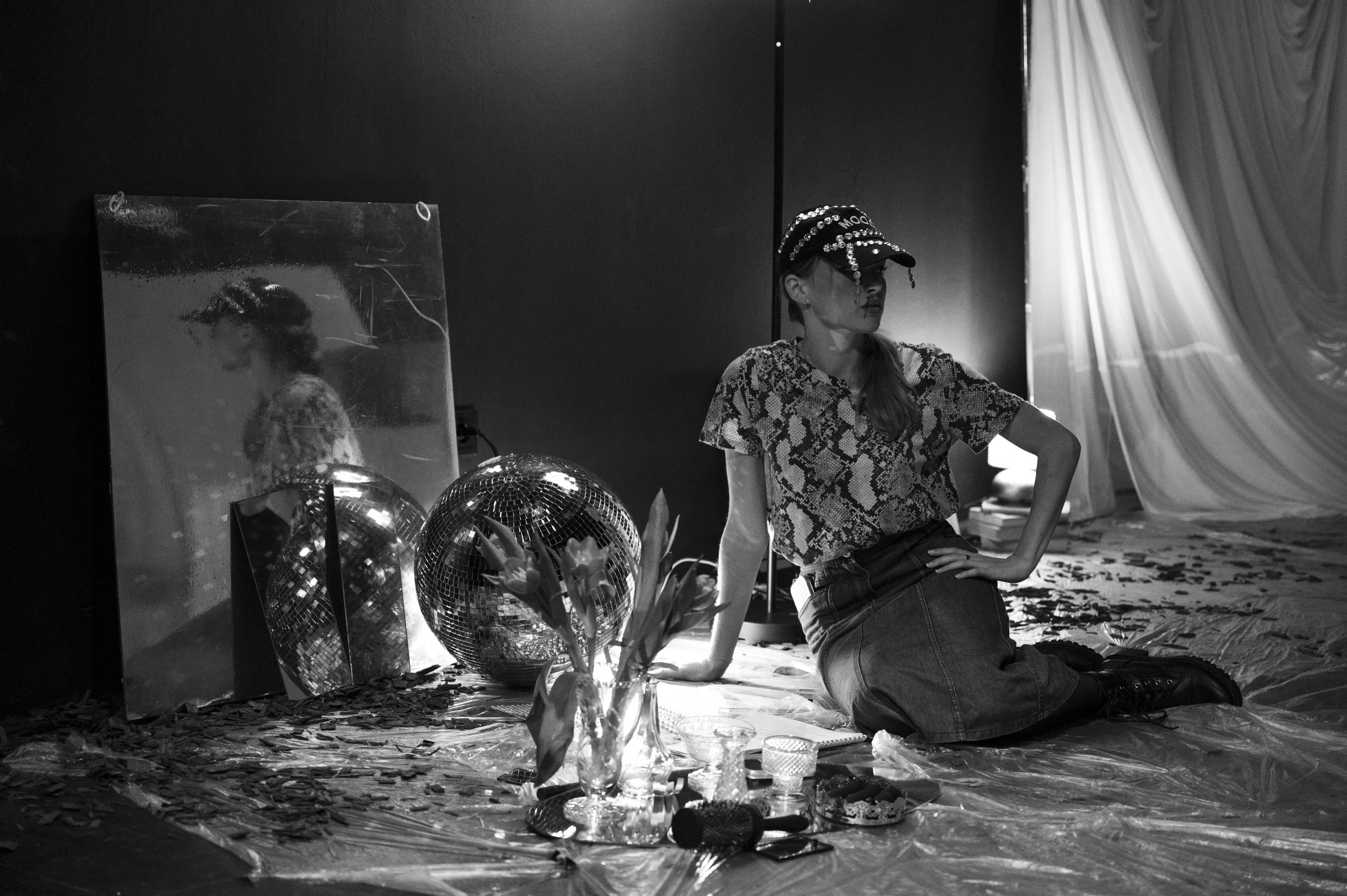 A woman wearing a patterned shirt, skirt, and cap sits on the floor in a room with dark walls. The floor is covered with plastic, and there are decorative items including a mirror, a disco ball, flowers, and small objects in front of her. A mirror reflects her profile and some of the objects.