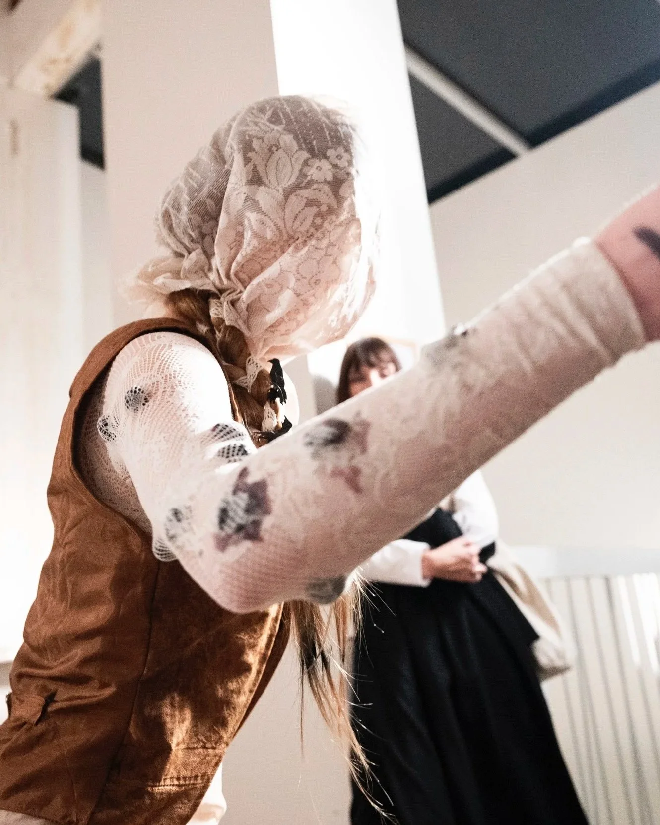 Person wearing a lace headscarf, lace long-sleeve shirt, and a brown vest, reaching out with one arm. Another person standing in the background observing.