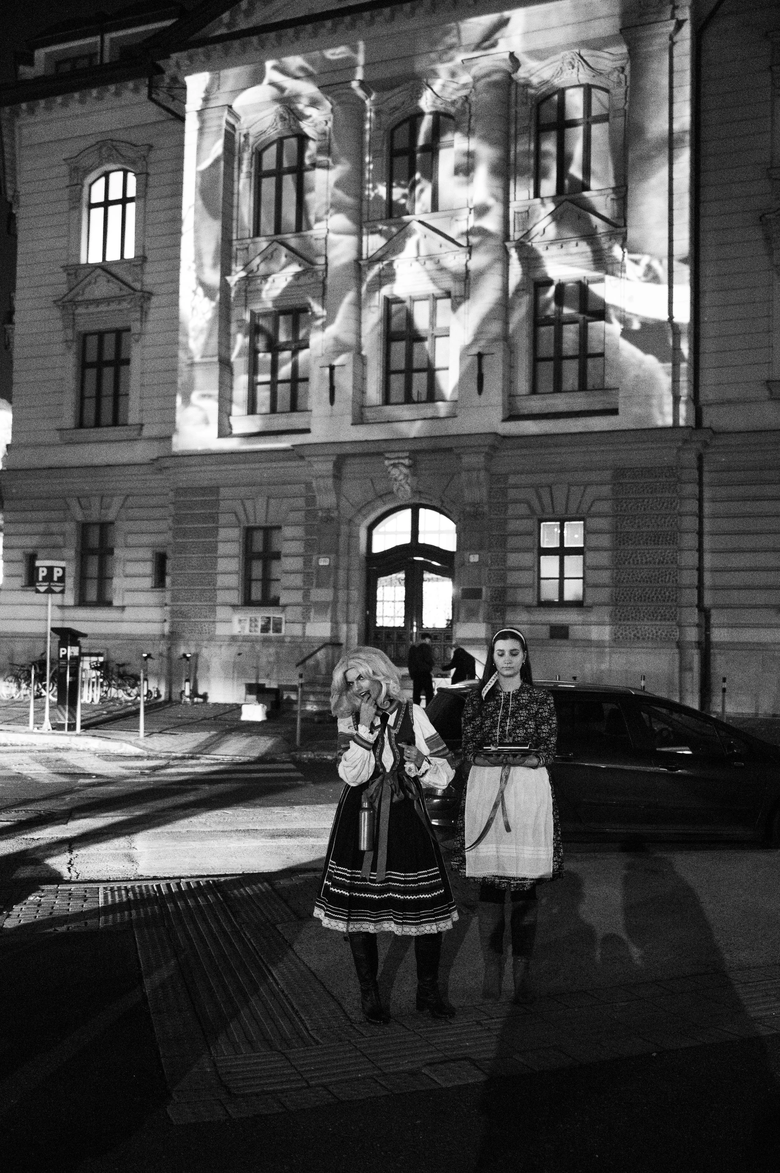 Black and white street scene at night with two women in traditional dresses standing near a car in front of a historic building. A projection of a child's face and hands is displayed on the building's facade.