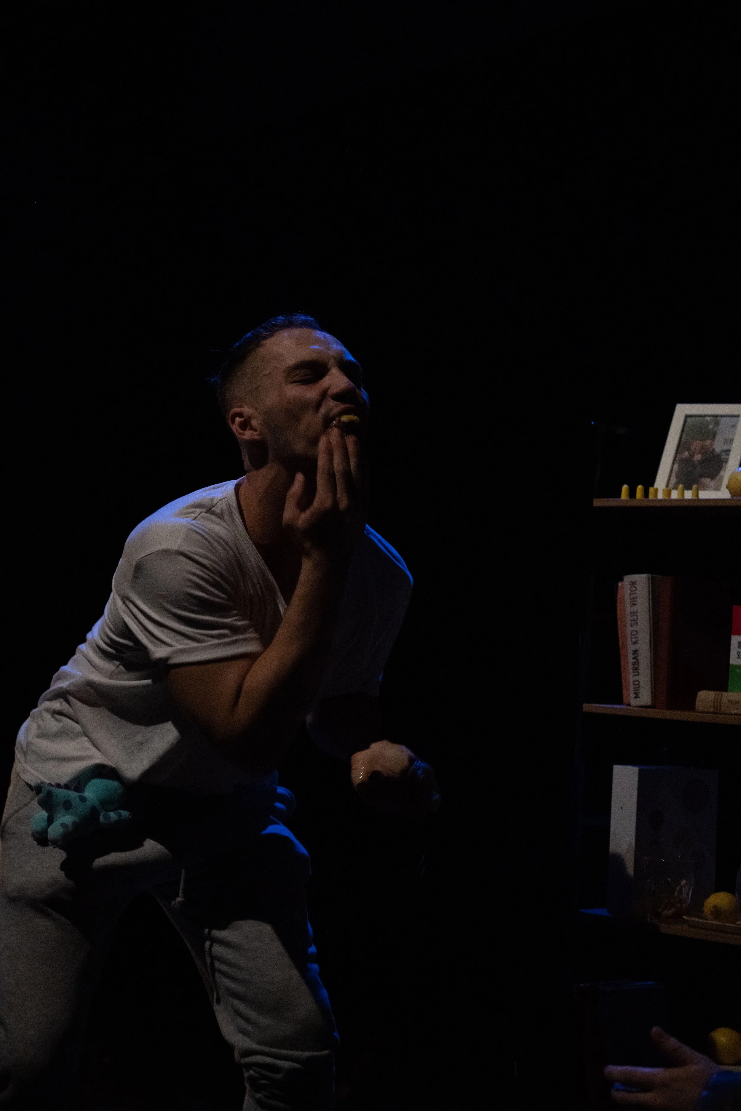 Person with short hair, wearing a white t-shirt and gray pants, sitting in a dark room with a small bookshelf visible in the background, appearing to be in distress or pain.