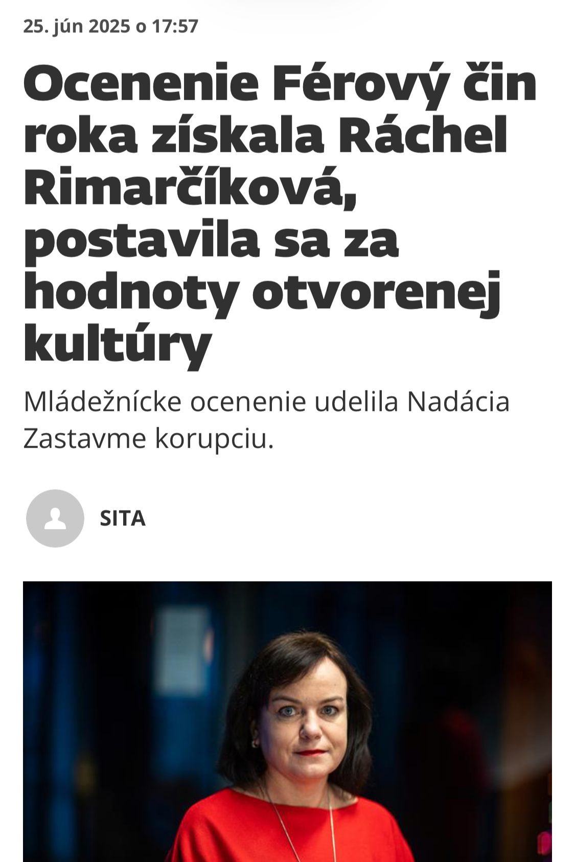 A woman with short dark hair wearing a red top and gold necklace, standing indoors with a blurry background.