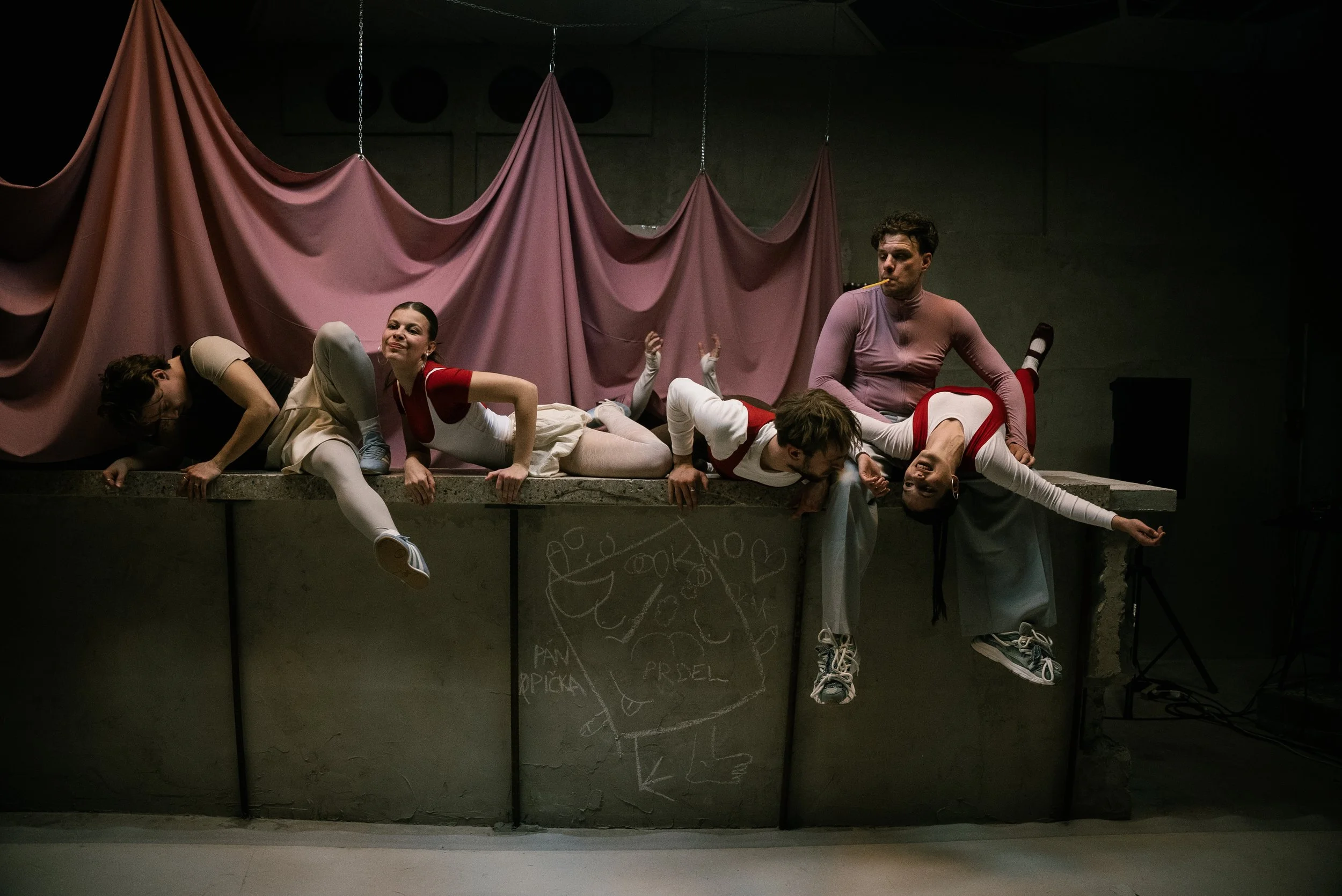 A theatrical performance with six actors on a stage, lying on a long concrete platform with a pink fabric backdrop. The actors are in various casual poses, some smiling, one smoking a cigarette, and a mix of relaxed and expressive expressions.