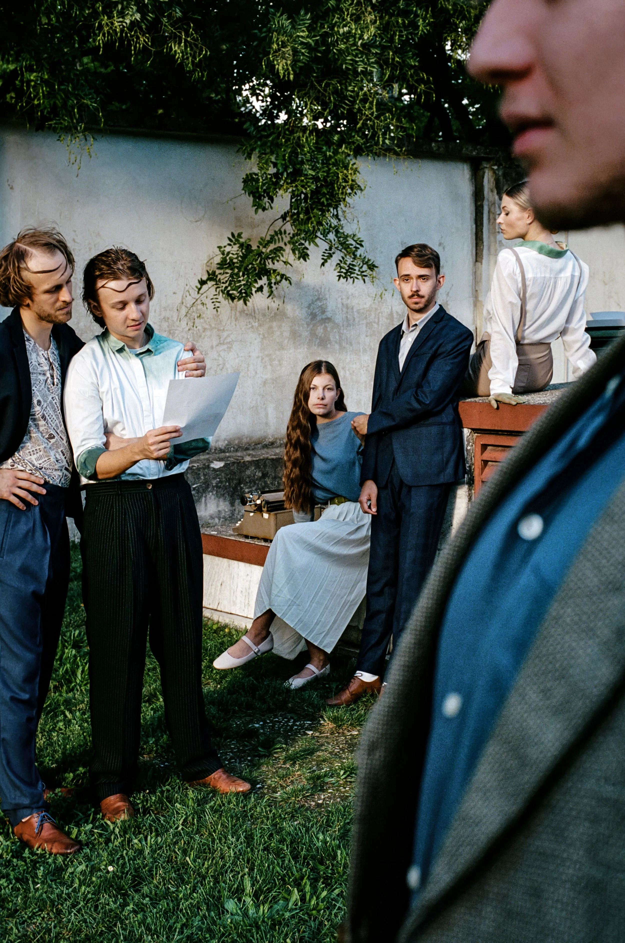 Group of six people outdoors near a wall, some engaged in reading or looking at papers, with one seated woman and a man in a suit in the center, and others standing or sitting nearby.