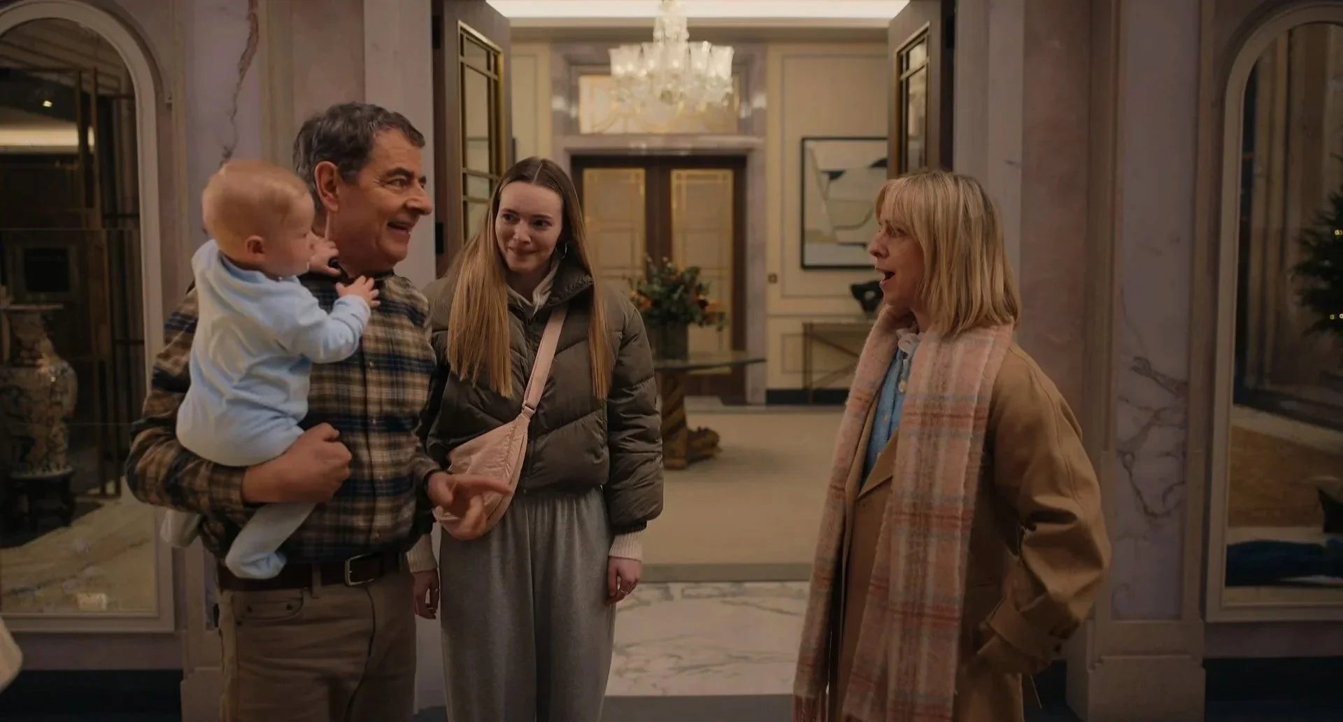 A man holding a baby and two women standing and talking inside a luxurious house with marble floors, chandeliers, and artwork.