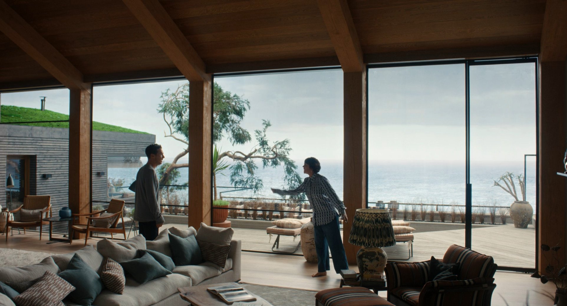 A man and a woman are having a conversation in a modern living room with large glass windows overlooking the ocean. The woman is leaning forward and gesturing towards the man, who is standing and listening. The room has wooden beams, a sofa with pillows, and various chairs and lamps, with outdoor furniture visible on a balcony.