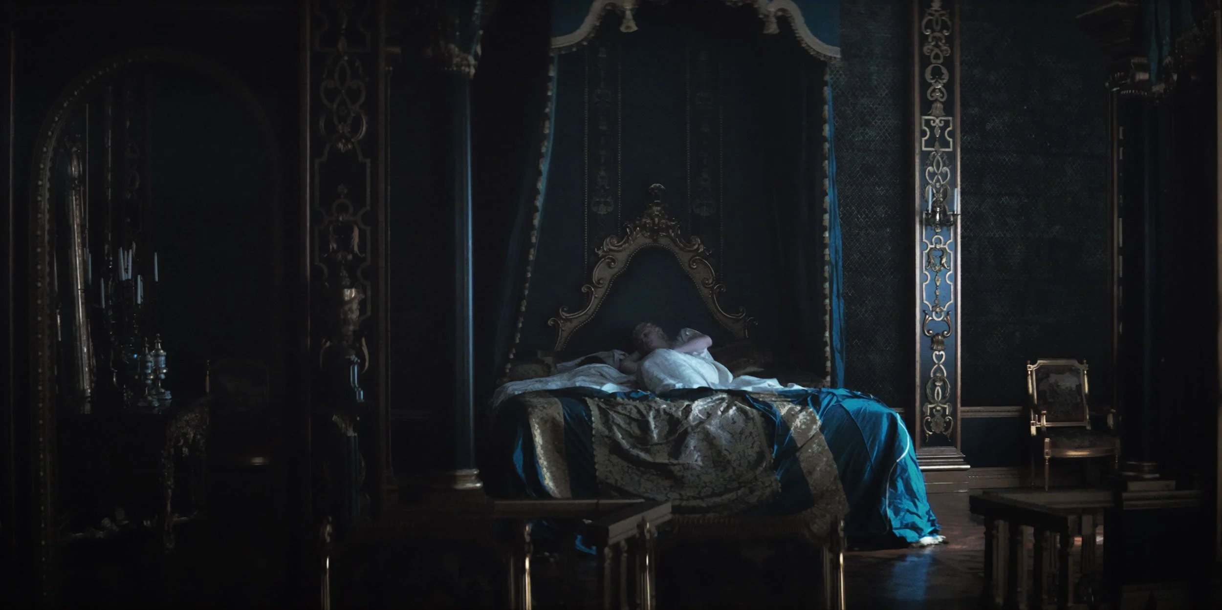 A person lying on an ornate bed in a dark, lavishly decorated room with gold accents and curtains.