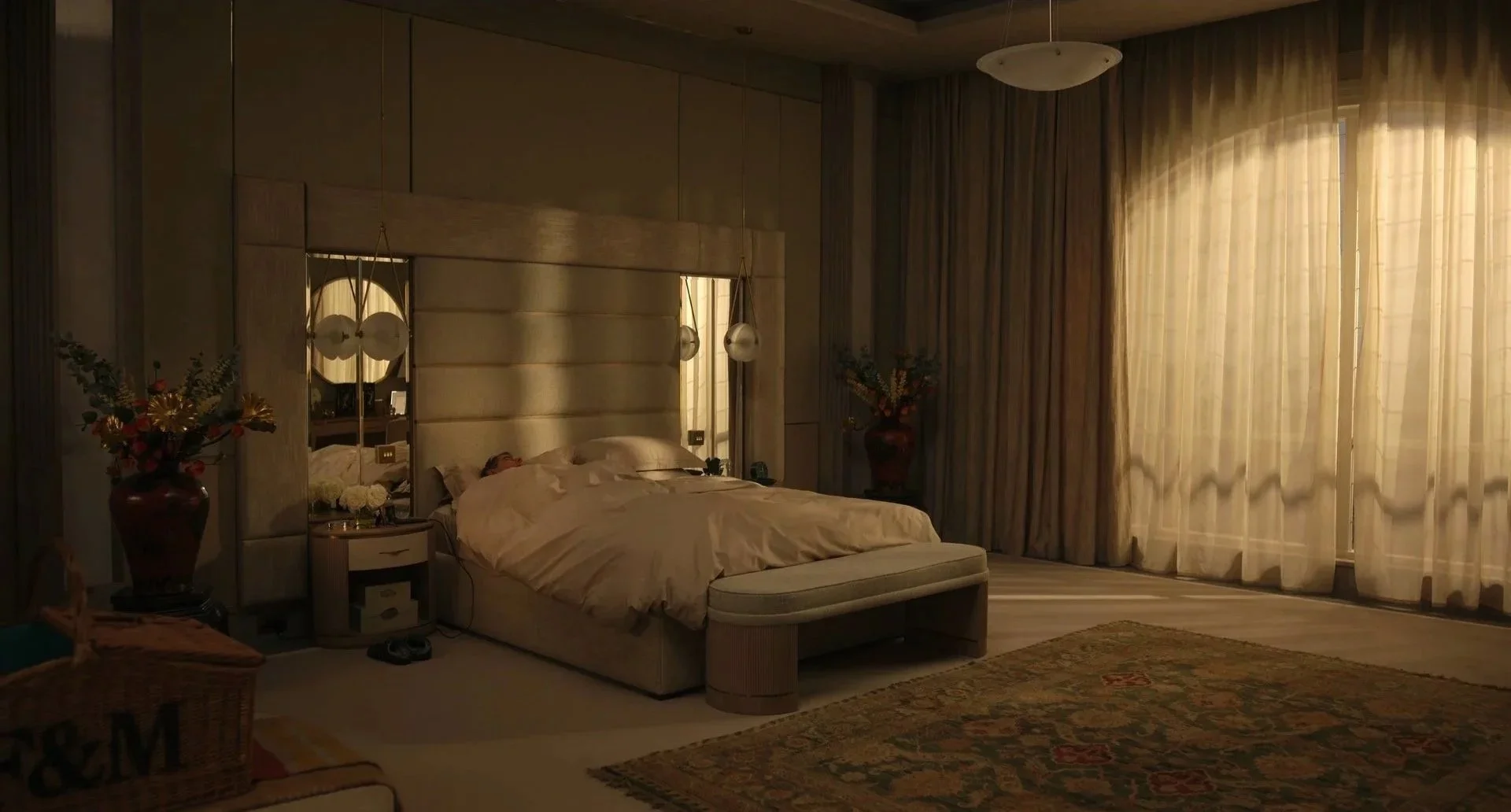 A bedroom with a large bed, person sleeping, gold and beige color scheme, large window with curtains, nightstand, floral arrangements, and soft lighting.