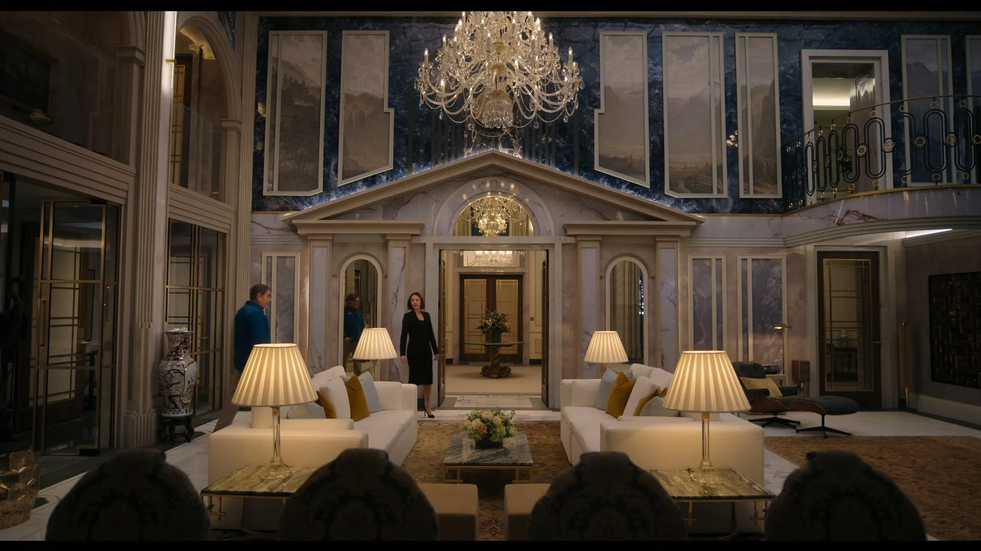 Elegant hotel lobby with chandeliers, marble walls, and symmetrical white sofas with gold accent pillows, with three people speaking near the entrance.