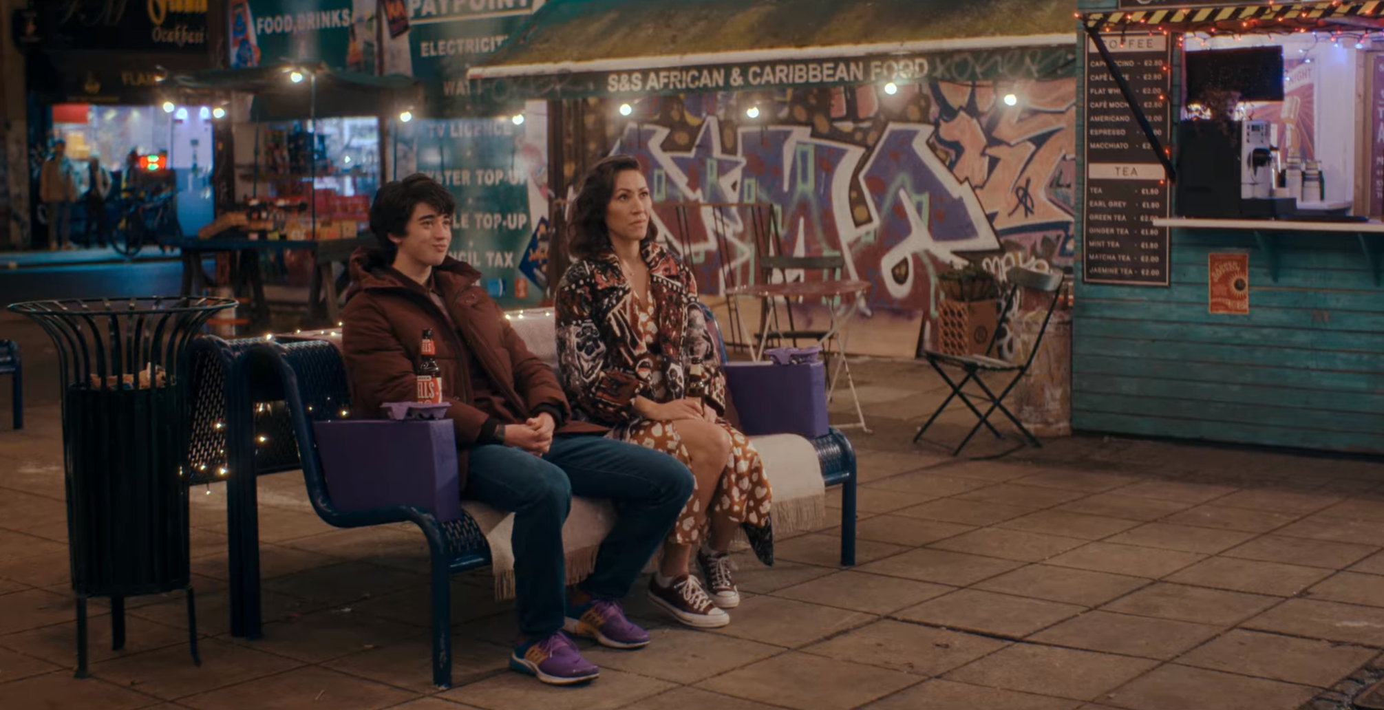 Two people sitting on a bench at a nighttime outdoor food market, with stalls and graffiti in the background.