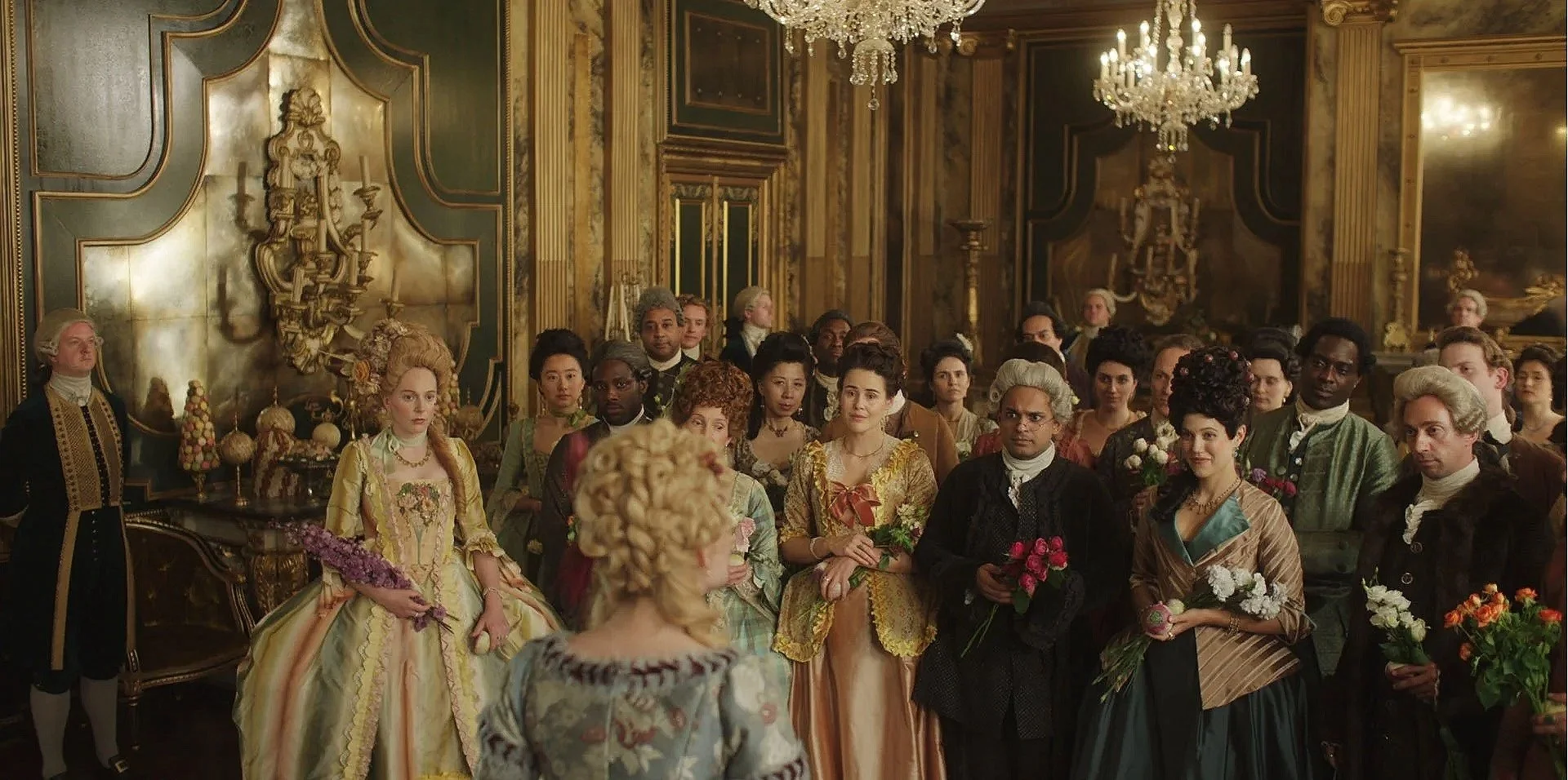 Group of people dressed in 18th-century period clothing at a formal gathering in an ornate, gilded hall with chandeliers and gold decor.