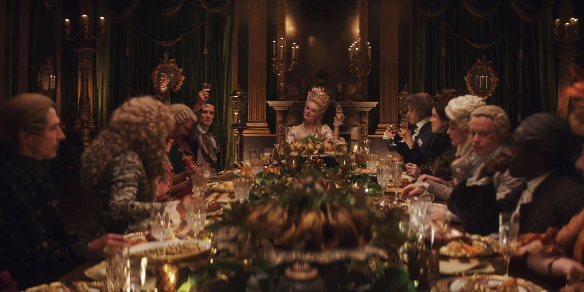 A lavish dinner party with elegantly dressed guests seated around a long, ornate table in a luxurious, gold-embellished room. The woman at the head holds a drink and smiles, surrounded by a mix of men and women in historical costumes.