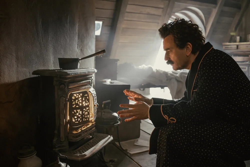 A man with curly hair and a mustache in pajamas is looking intently at a small vintage stove in a dimly lit attic room with wooden walls and sloped ceiling, sunlight filtering through an arched window.