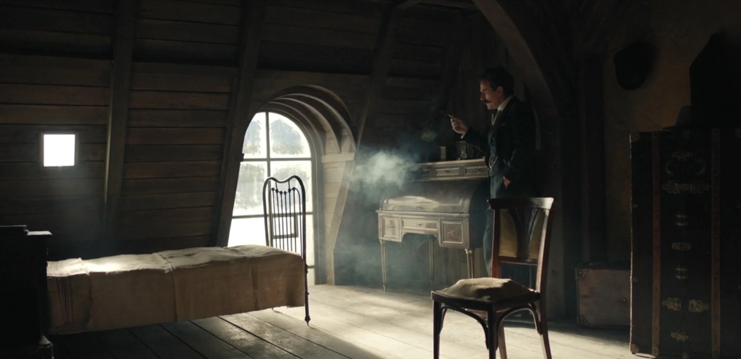 A man in a dark suit standing in a rustic attic room, smoking a cigarette, with light coming through a large arched window behind him. The room features wooden walls, a vintage bed, and an ornate white desk.
