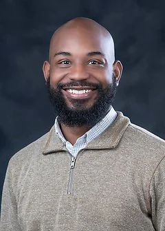 A smiling man with a beard and shaved head wearing a light gray zip-up sweater, standing against a dark background.