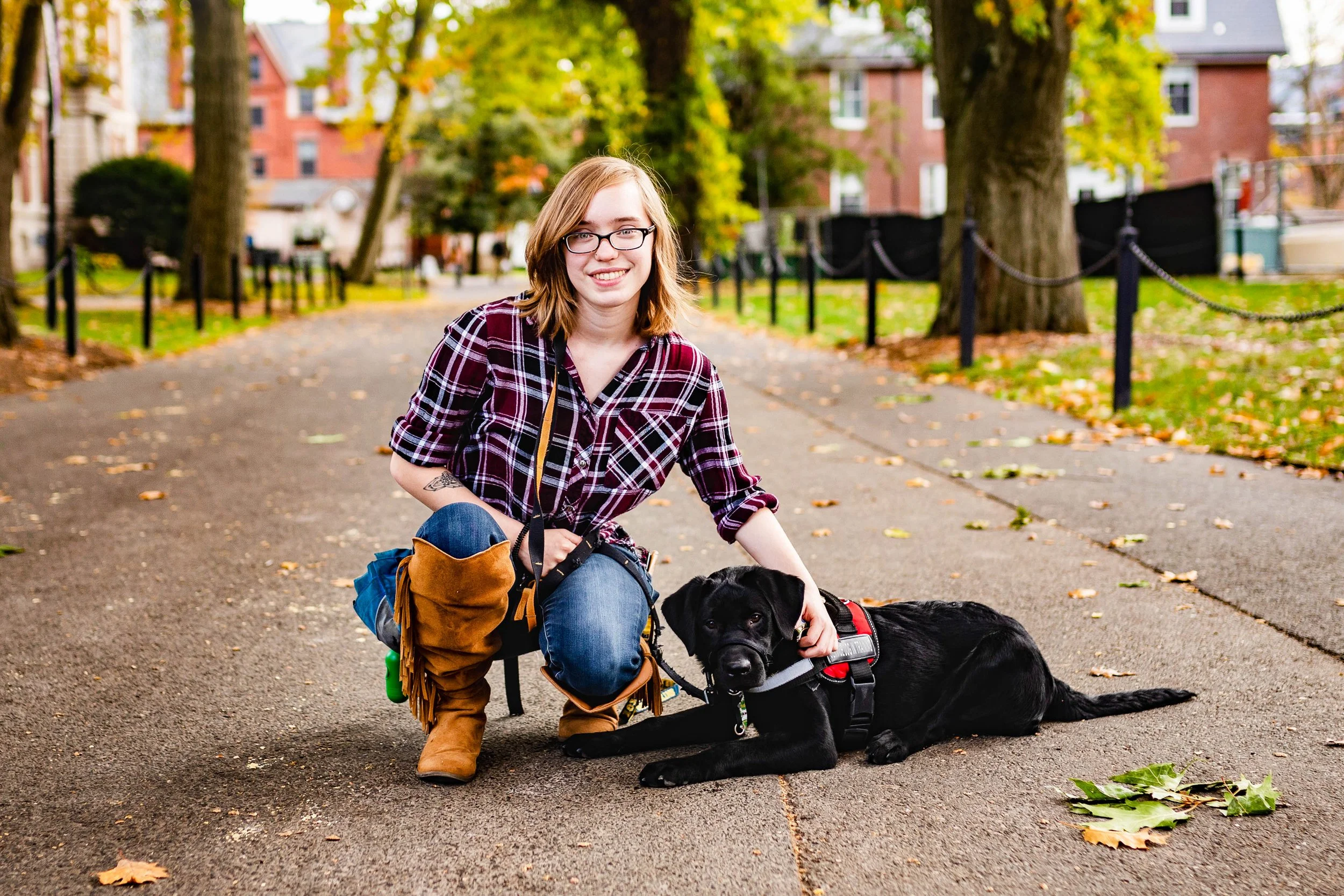 A woman wearing glasses and a red plaid shirt kneels next to a black Labrador Retriever lying on the street in a park with autumn leaves and trees.