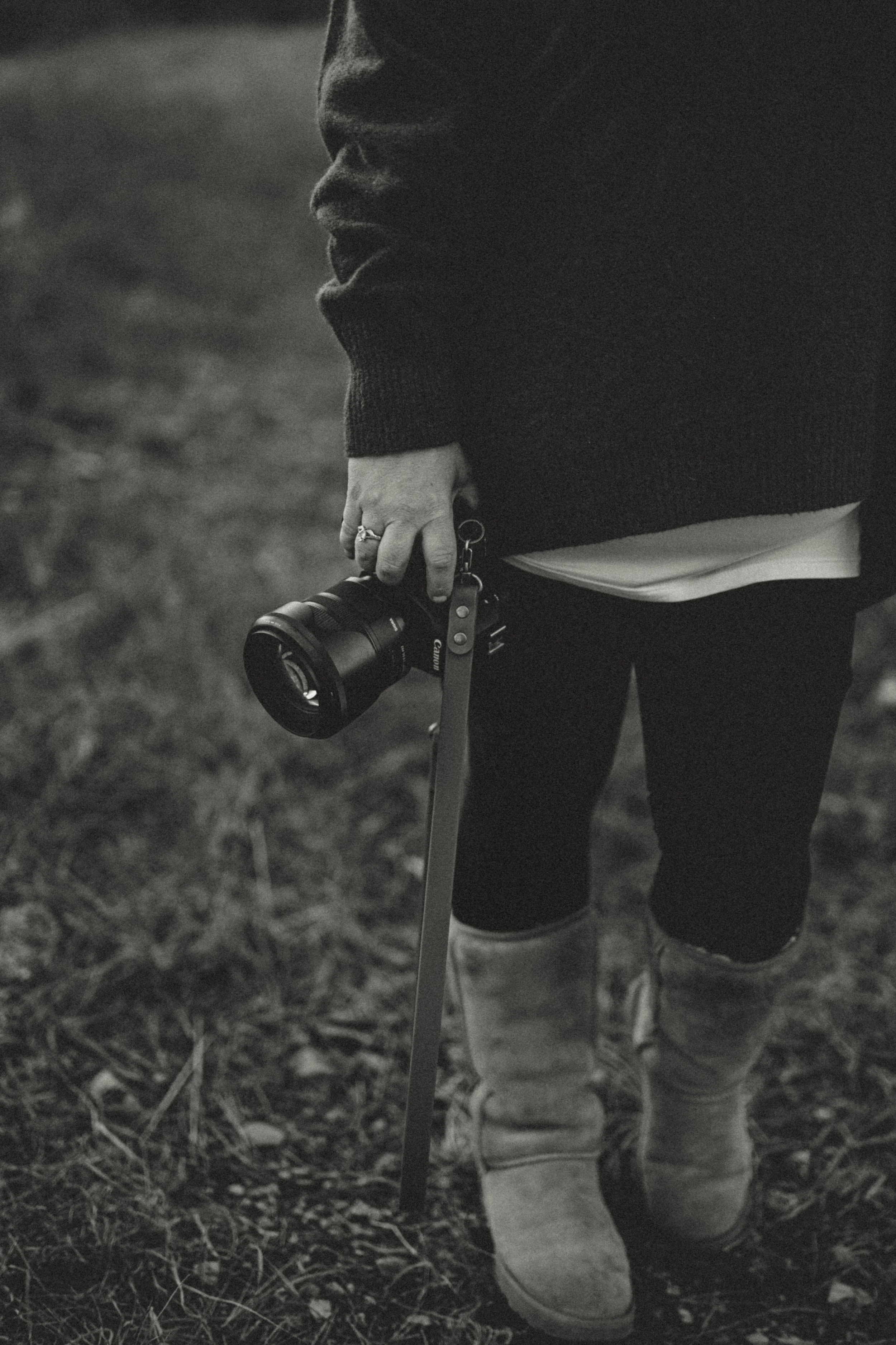 A person standing outdoors, holding a camera with a strap, dressed in a dark sweater, white shirt, dark pants, and light-colored boots.