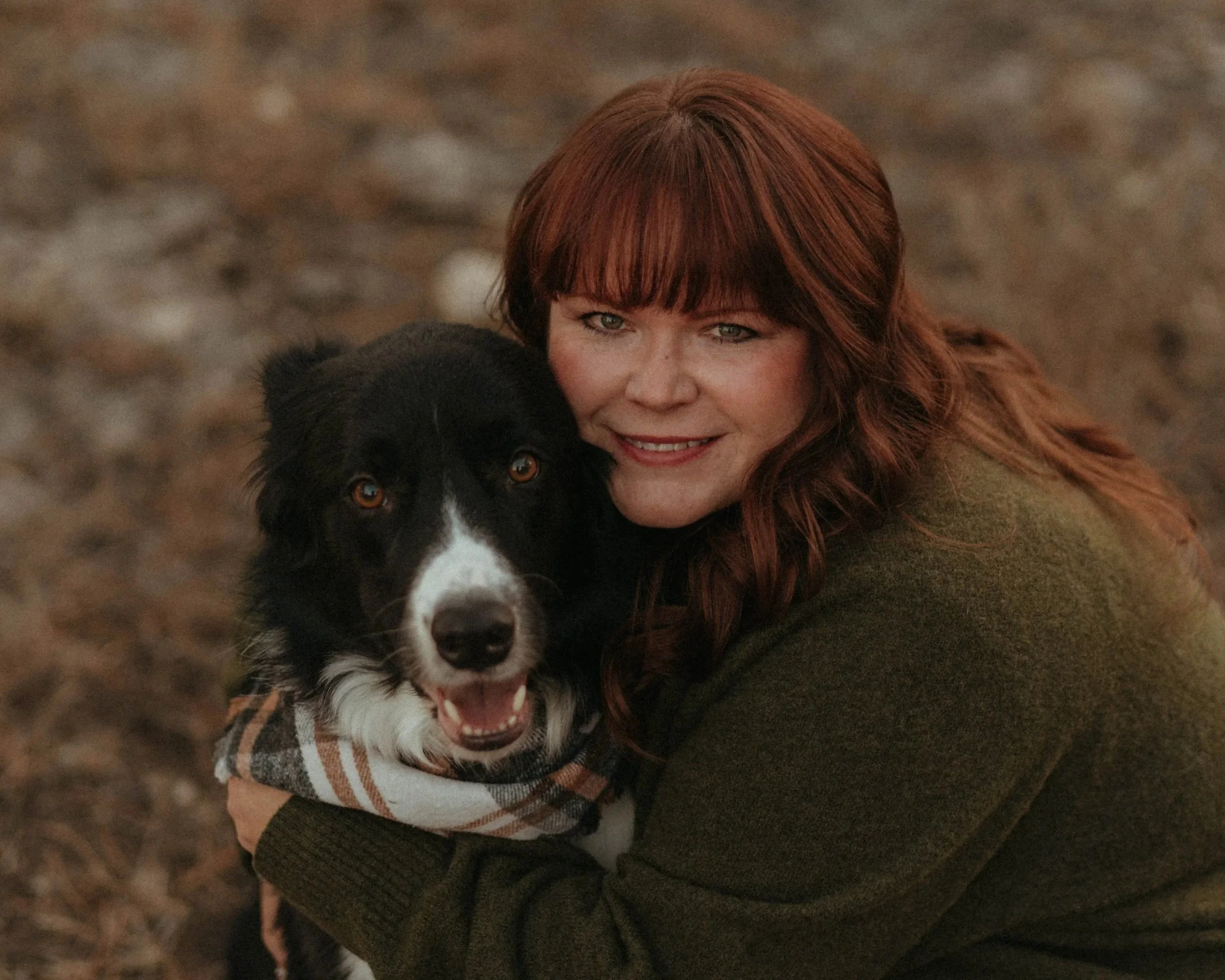 A woman with long red hair and bangs smiling while hugging a black and white Border Collie dog outside on a dirt ground.