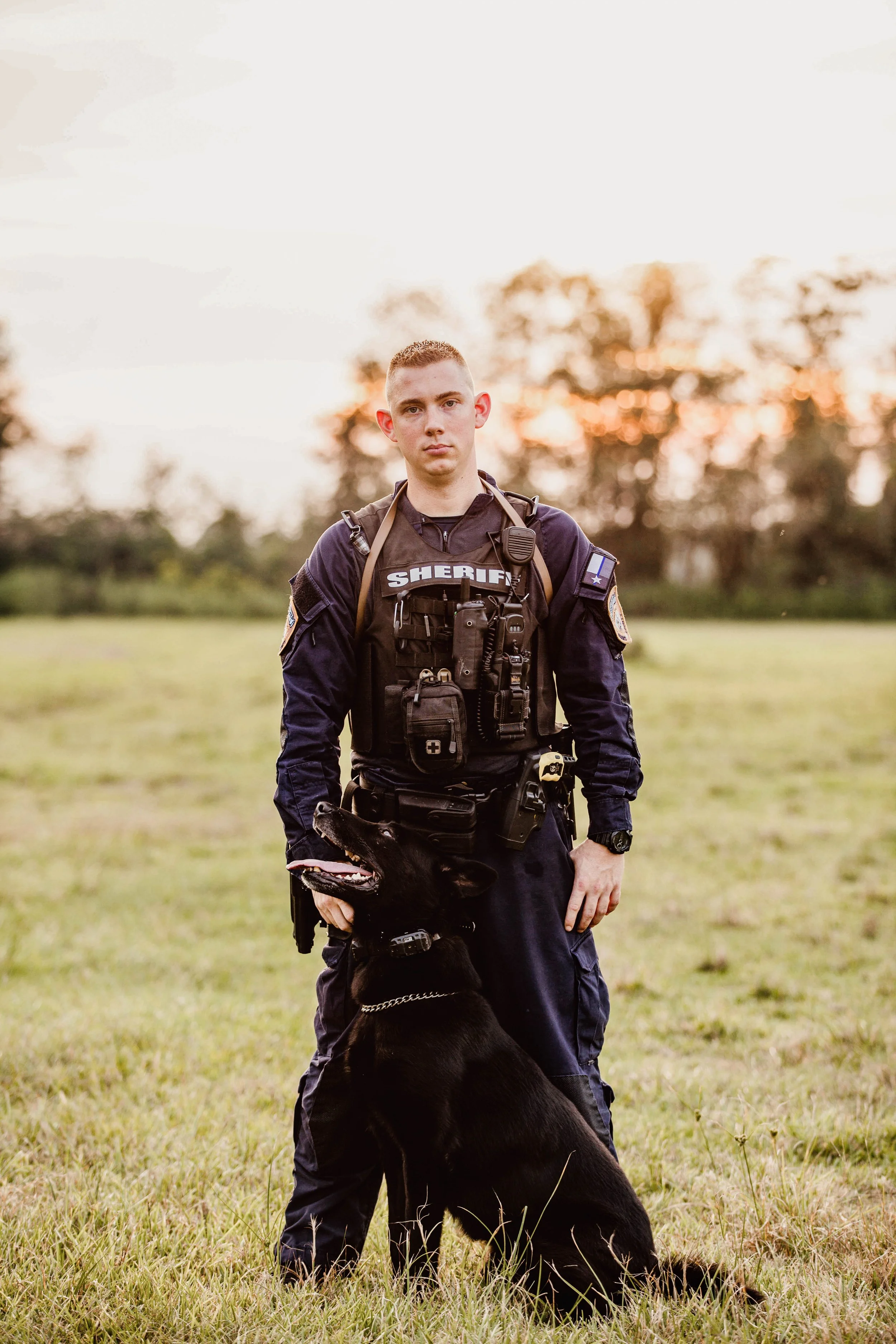 A young male sheriff's deputy in uniform standing outdoors at sunset with a black service dog.
