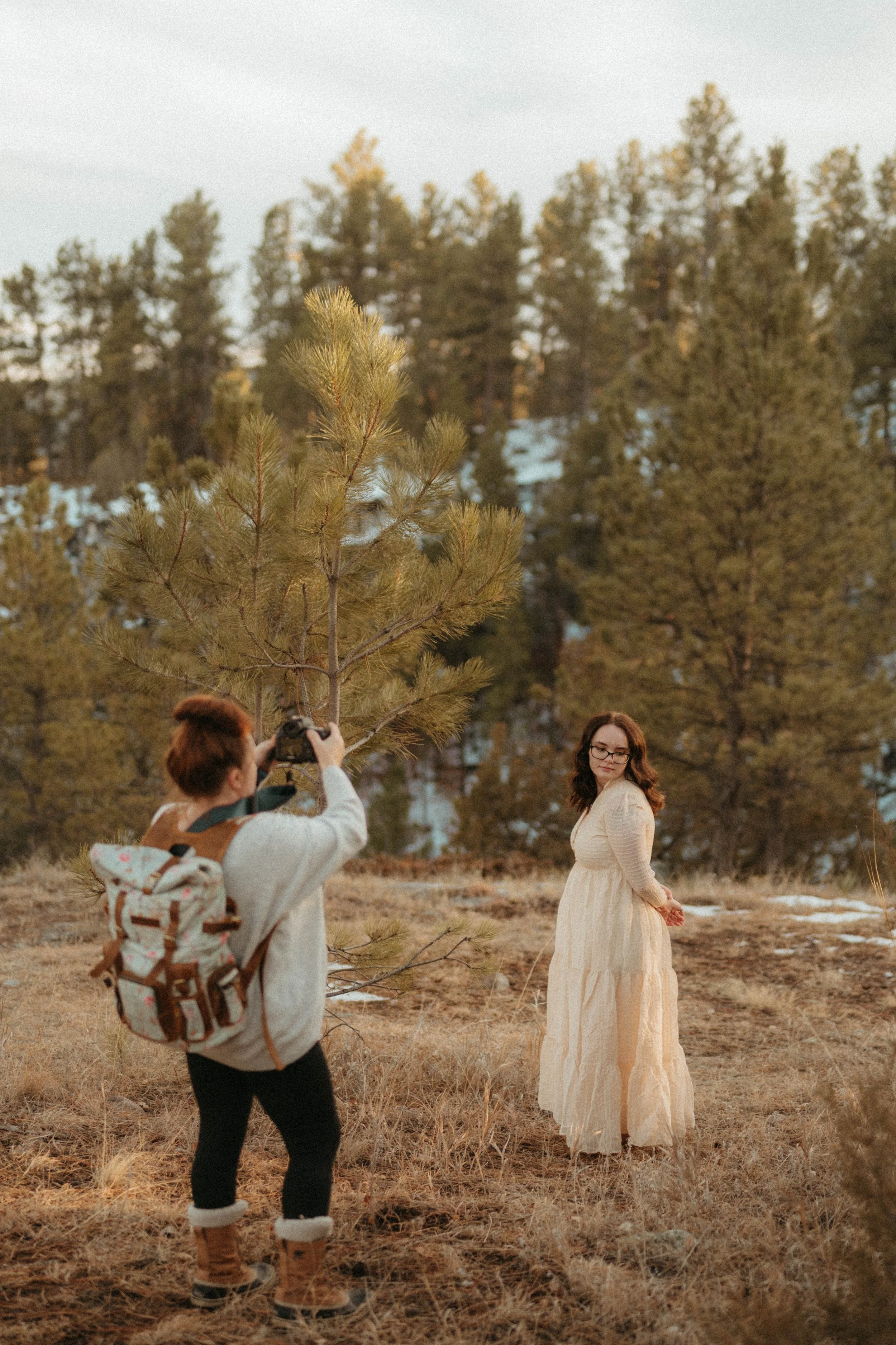A woman in a long cream dress poses in a wooded outdoor area while a person takes her photo with a camera.