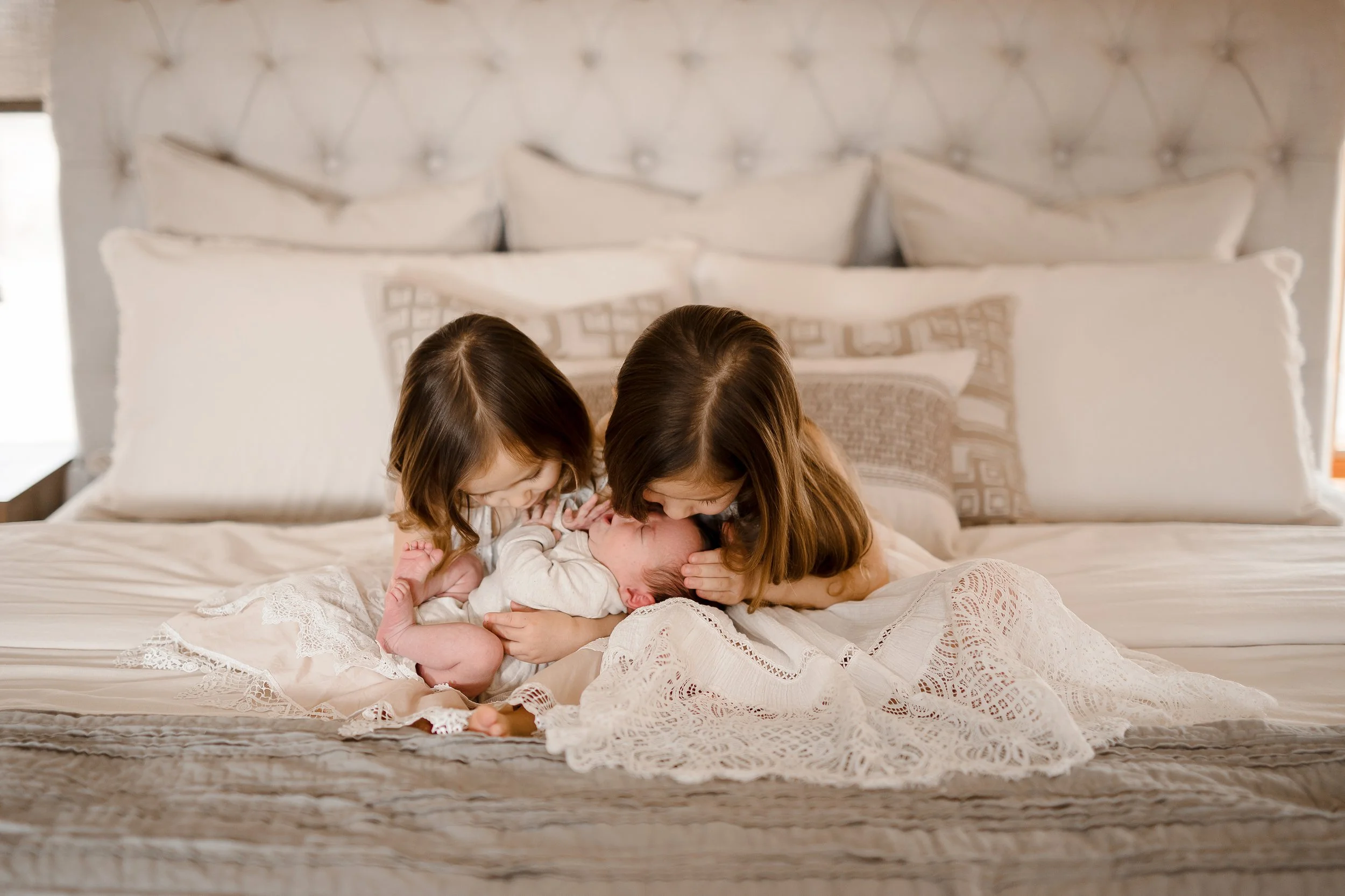 Two young girls kissing and cuddling a newborn baby on a bed in a cozy bedroom.