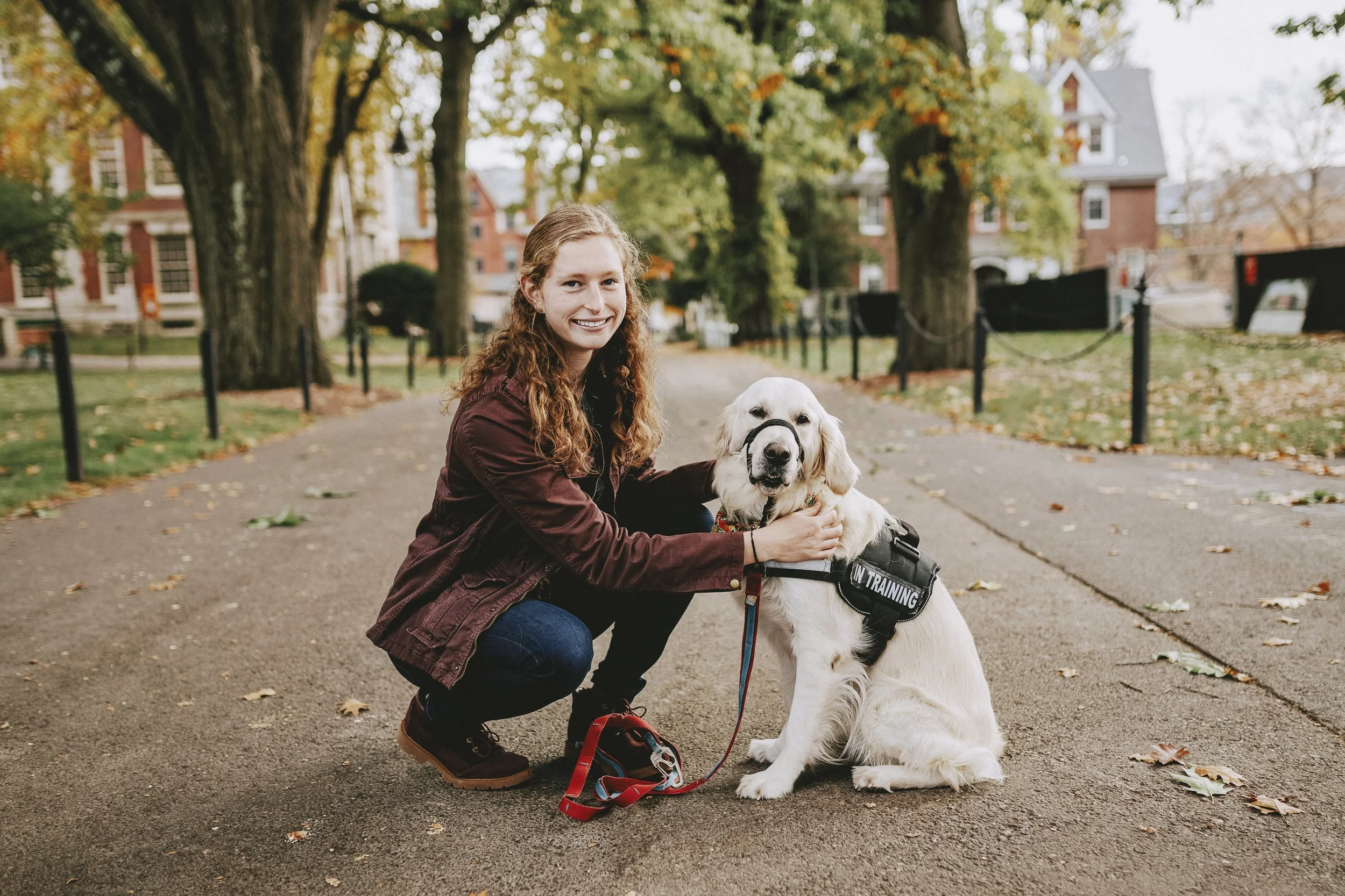 A young woman with curly red hair crouches down next to a guide dog in training, both smiling at the camera. The dog is sitting on a paved path in a park lined with trees and orange, yellow, and green autumn leaves.