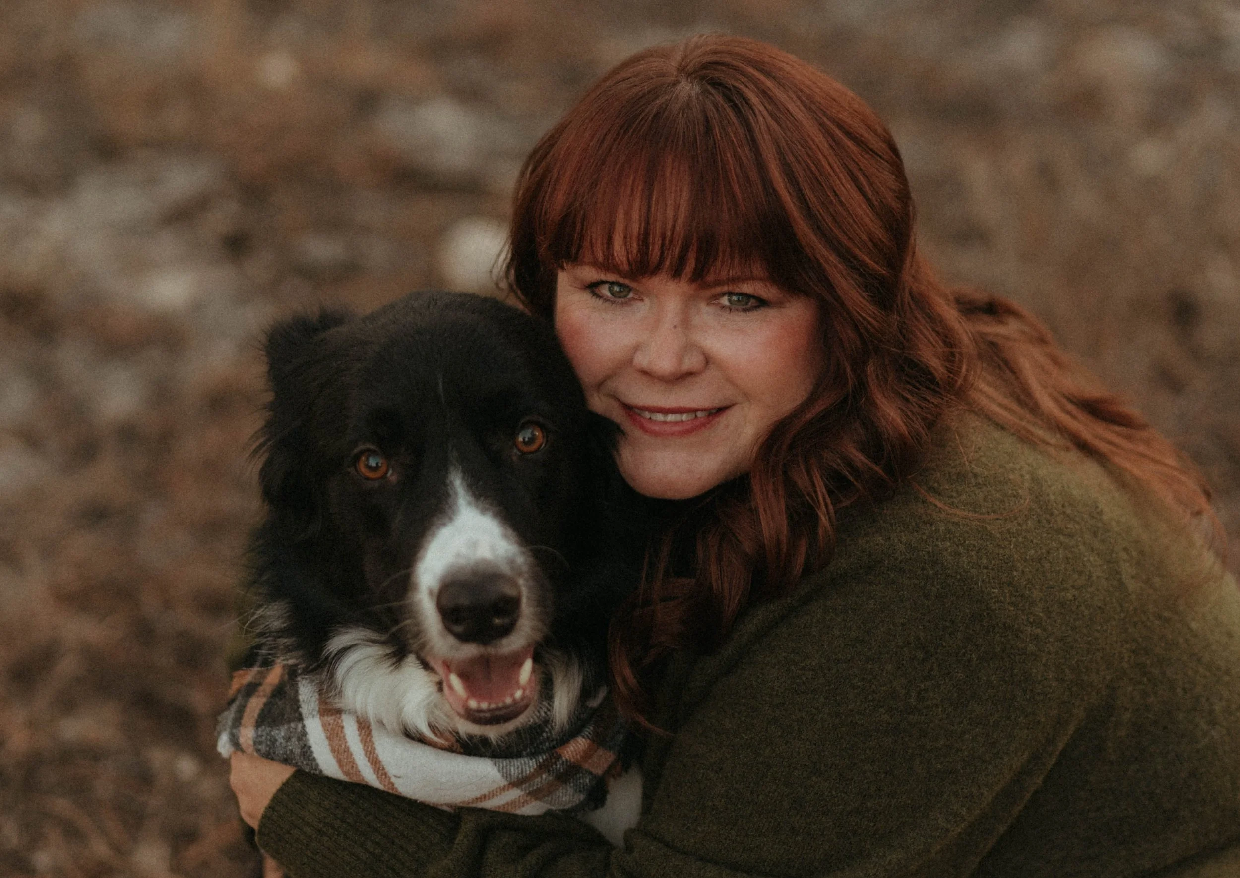 A woman with red hair smiling and hugging a black and white dog with a plaid bandana on its neck, outdoors on a dirt ground.