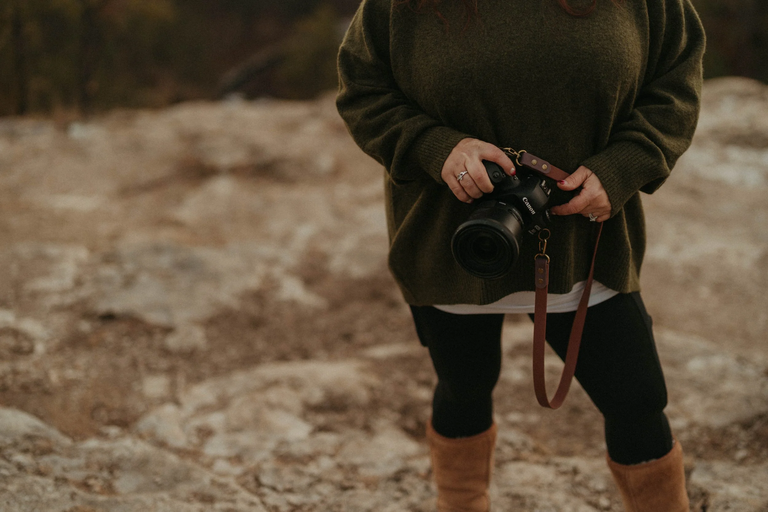 Person holding a Canon camera outdoors during sunset or dusk, wearing a green sweater and tan boots.