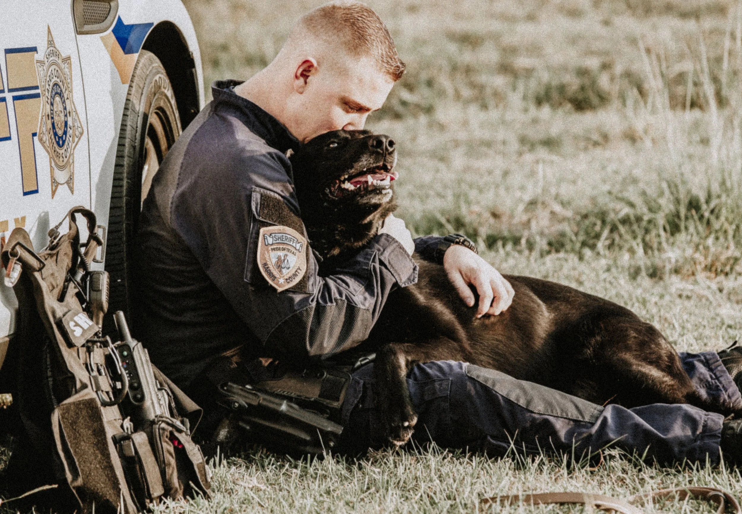 A sheriff's deputy sitting on the grass next to a police vehicle, hugging and comforting a black German Shepherd police dog that is lying on the ground. The deputy is wearing a uniform with an sheriff patch on the sleeve.