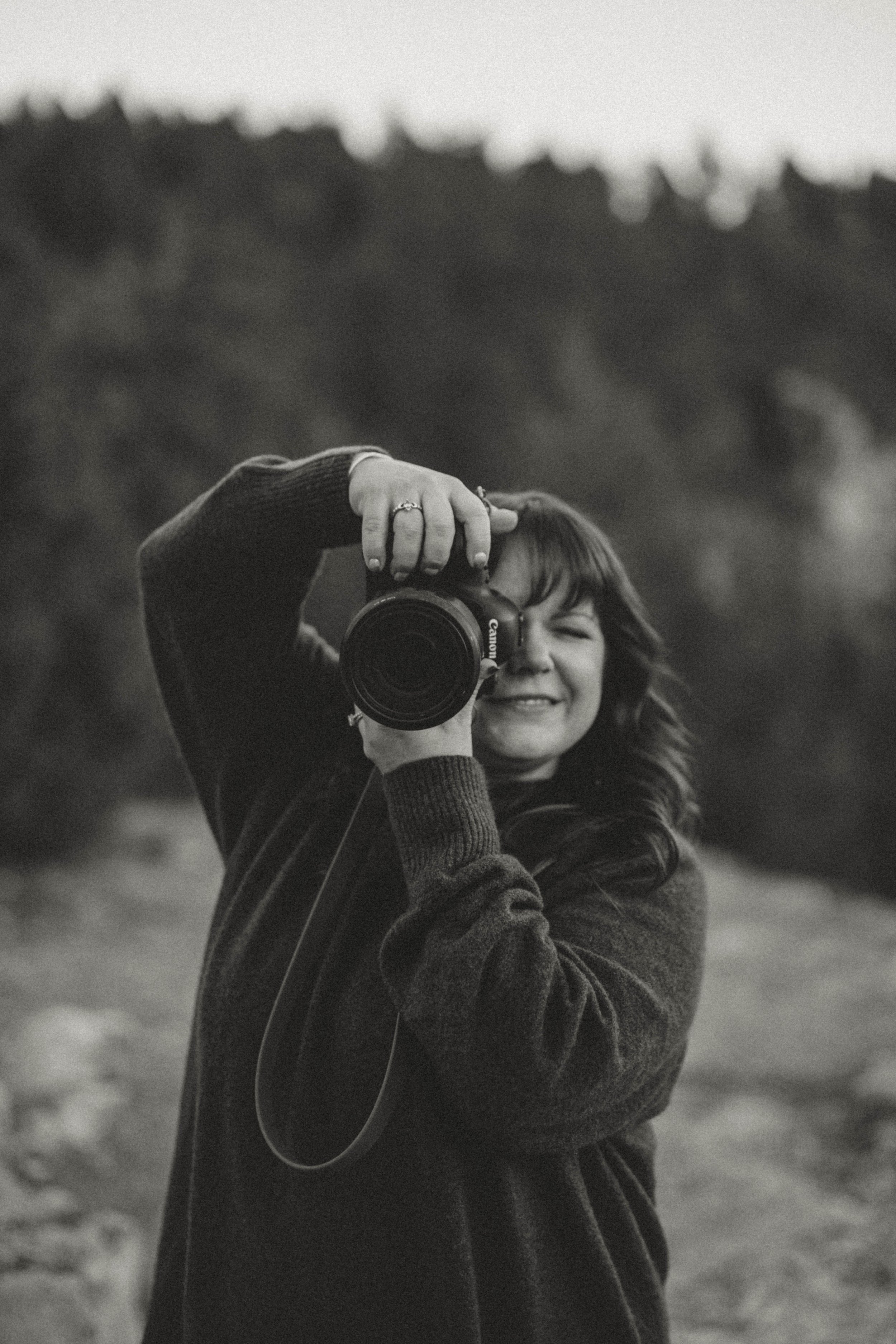 A woman with wavy hair taking a photo with a Canon camera outdoors, smiling with one eye closed.