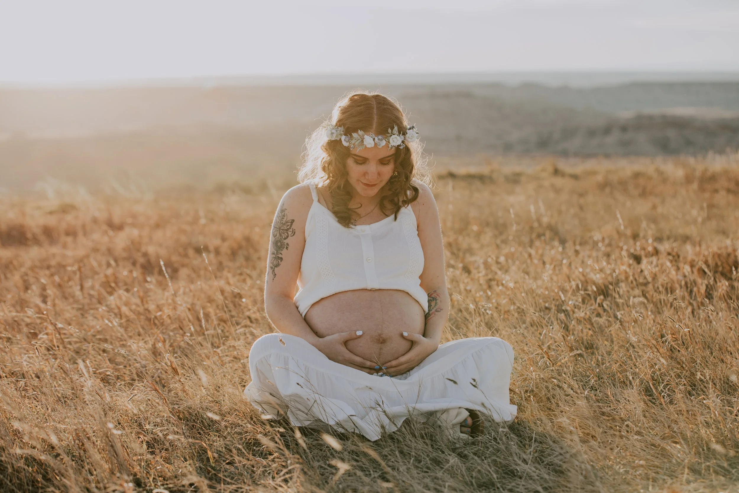 A pregnant woman sits cross-legged in a field of tall grass during sunset, wearing a white sleeveless top, white skirt, a floral headband, and showing tattoos on her arms.