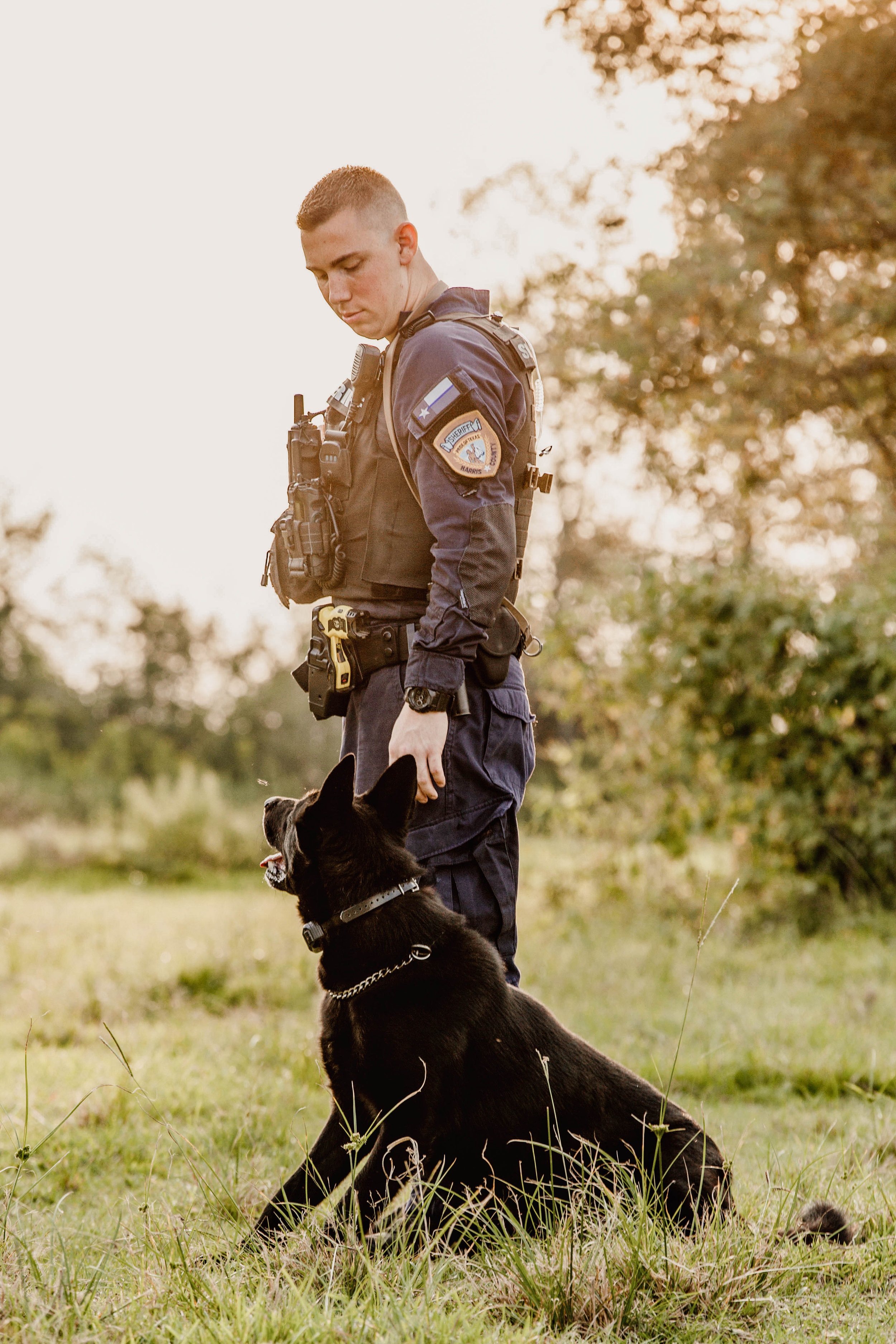 A police officer in uniform with a police patch on the sleeve, standing outdoors on grass with a black police dog sitting at his feet, during sunset or sunrise.