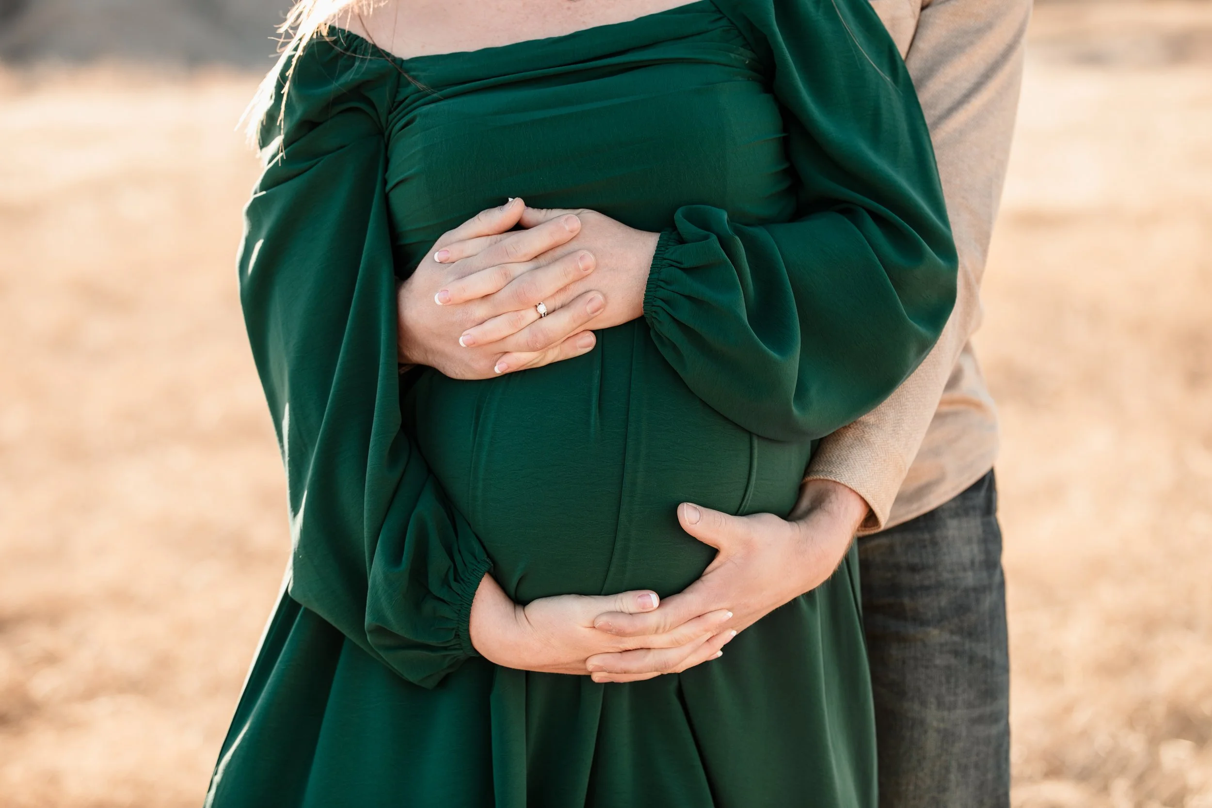 A pregnant woman in a green dress with hands on her belly, standing outdoors with a man behind her holding her.