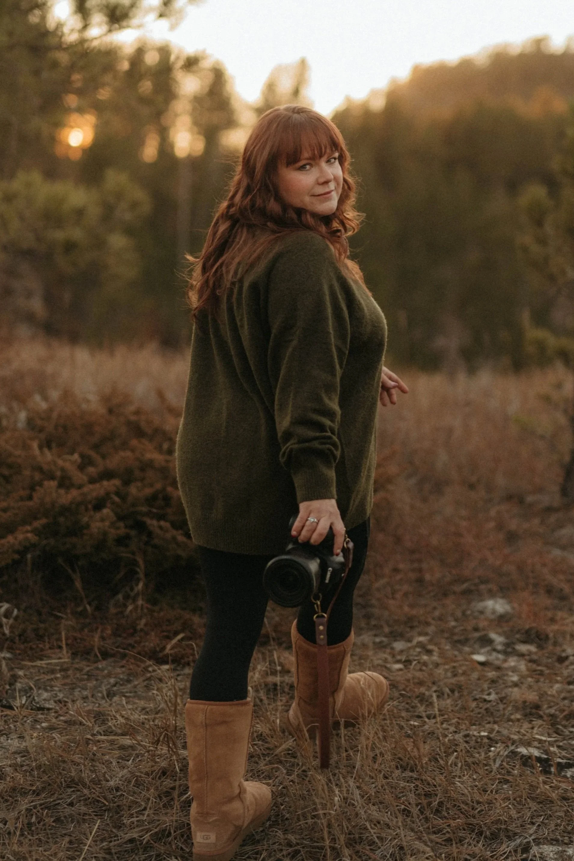 Woman standing outdoors in a field during sunset, holding a camera, wearing a green sweater, black leggings, and tan high-heeled boots.