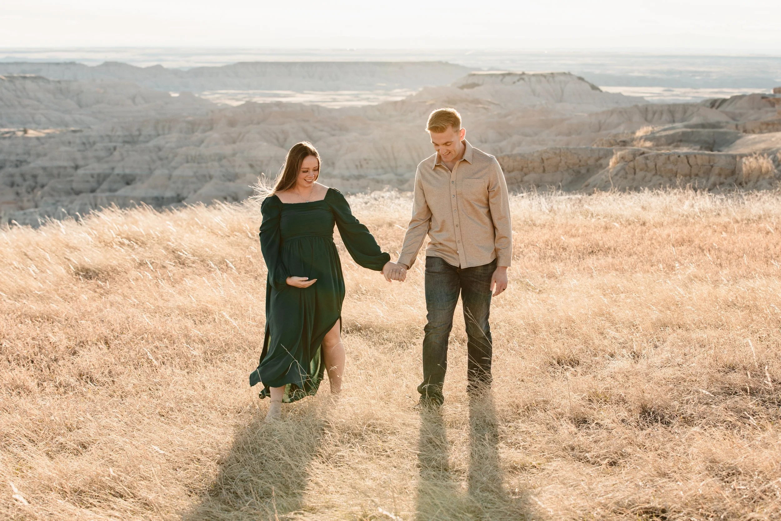 A pregnant woman and a man holding hands and walking in a field of tall, golden grass with a desert landscape in the background during sunset.