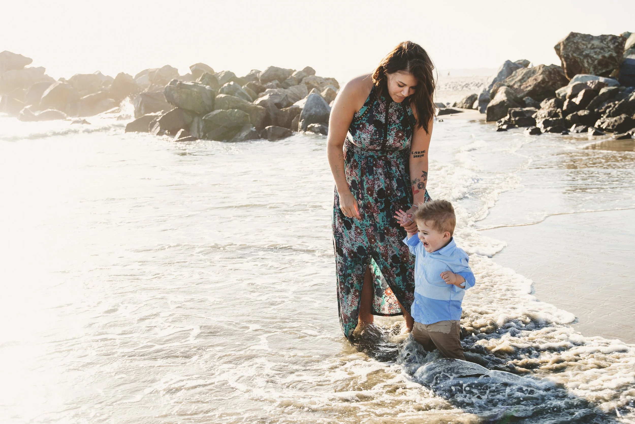 A woman and a young boy playing in the ocean at the beach near rocks, enjoying a sunny day.