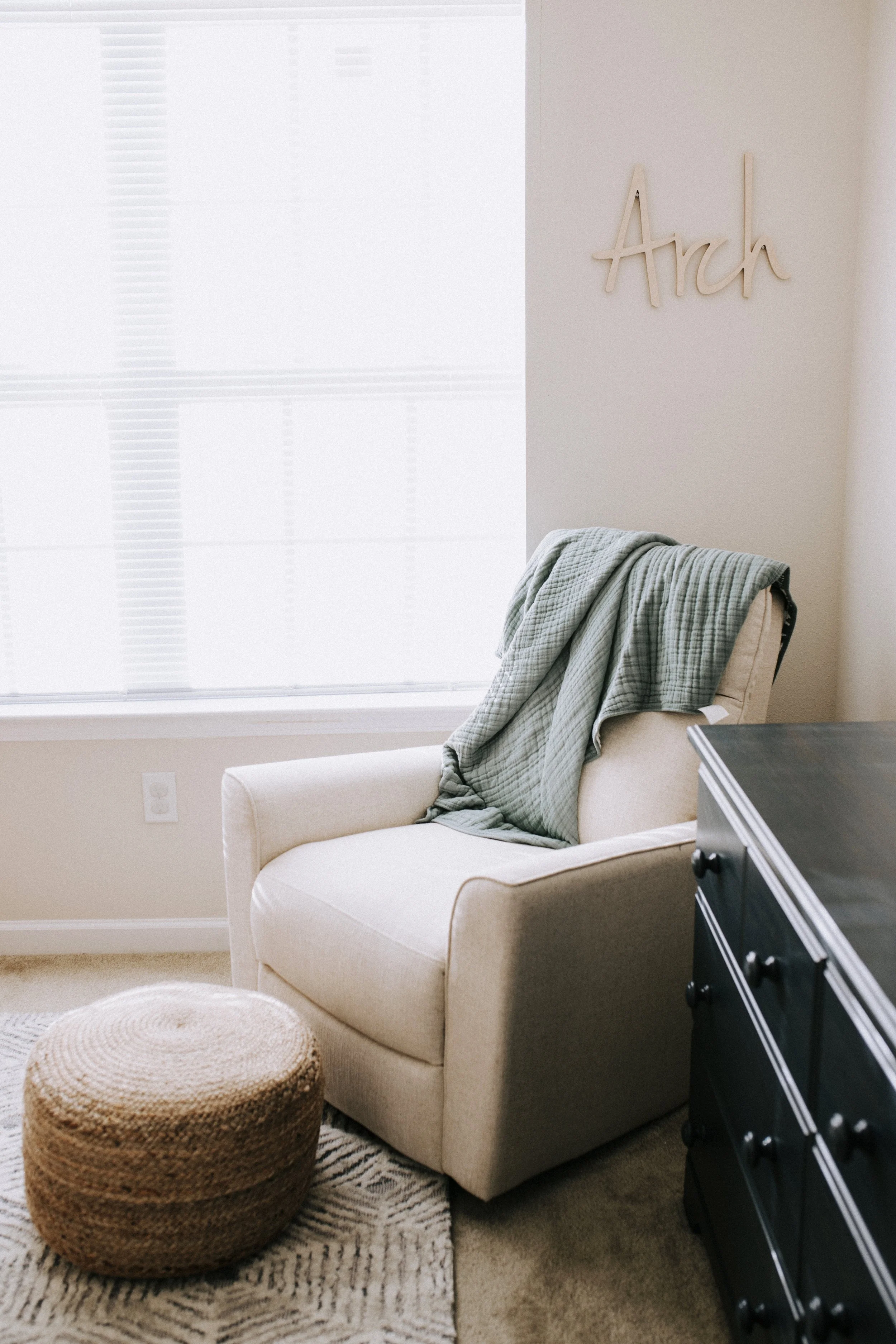 A cozy corner with a beige armchair draped with a green blanket, a woven pouf ottoman, a black dresser, and a wall decoration that says 'Arch' near a window with white blinds.