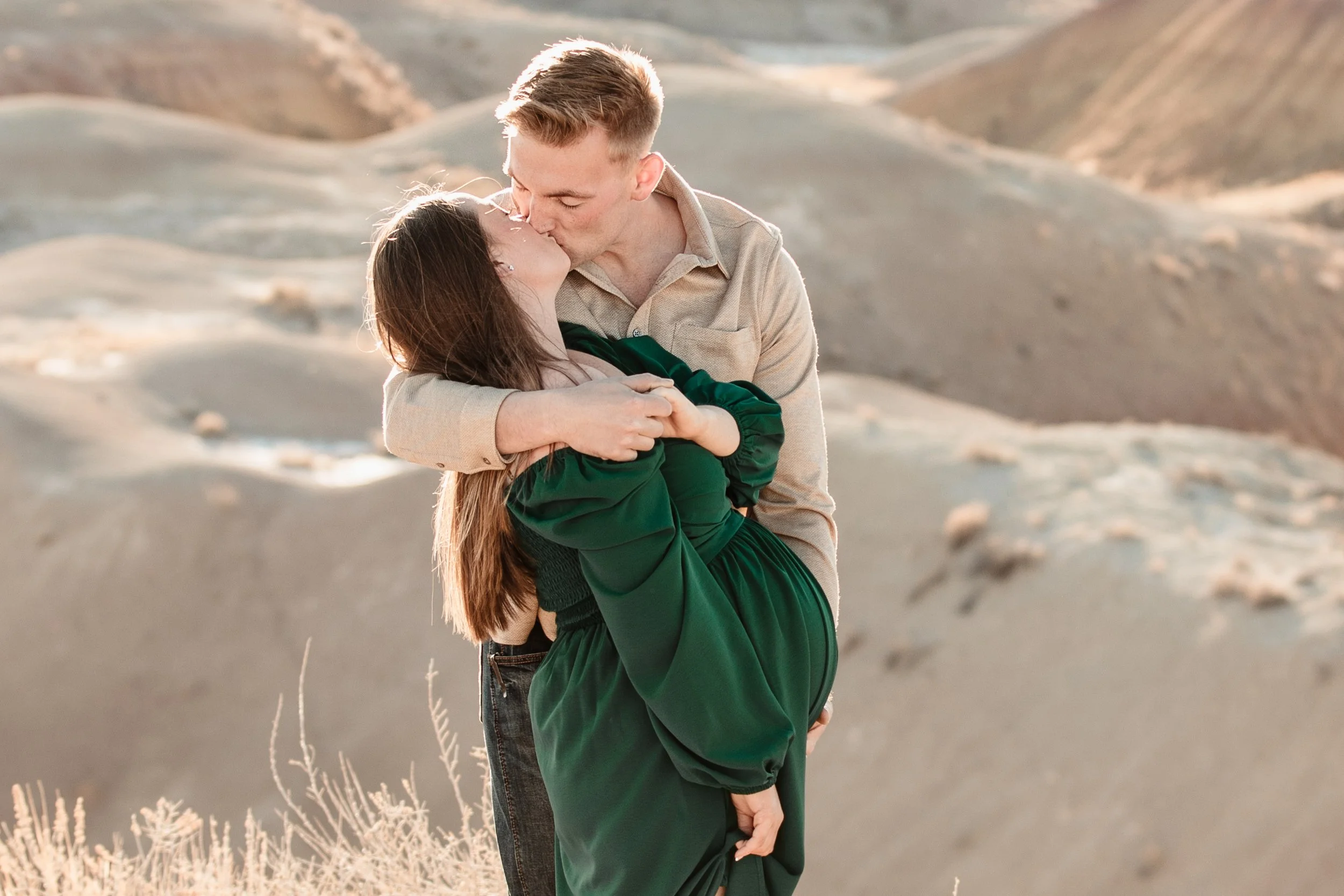 A couple kissing in a desert landscape during sunset. The man is wearing a beige shirt and has his arms around the woman, who is wearing a dark green dress.