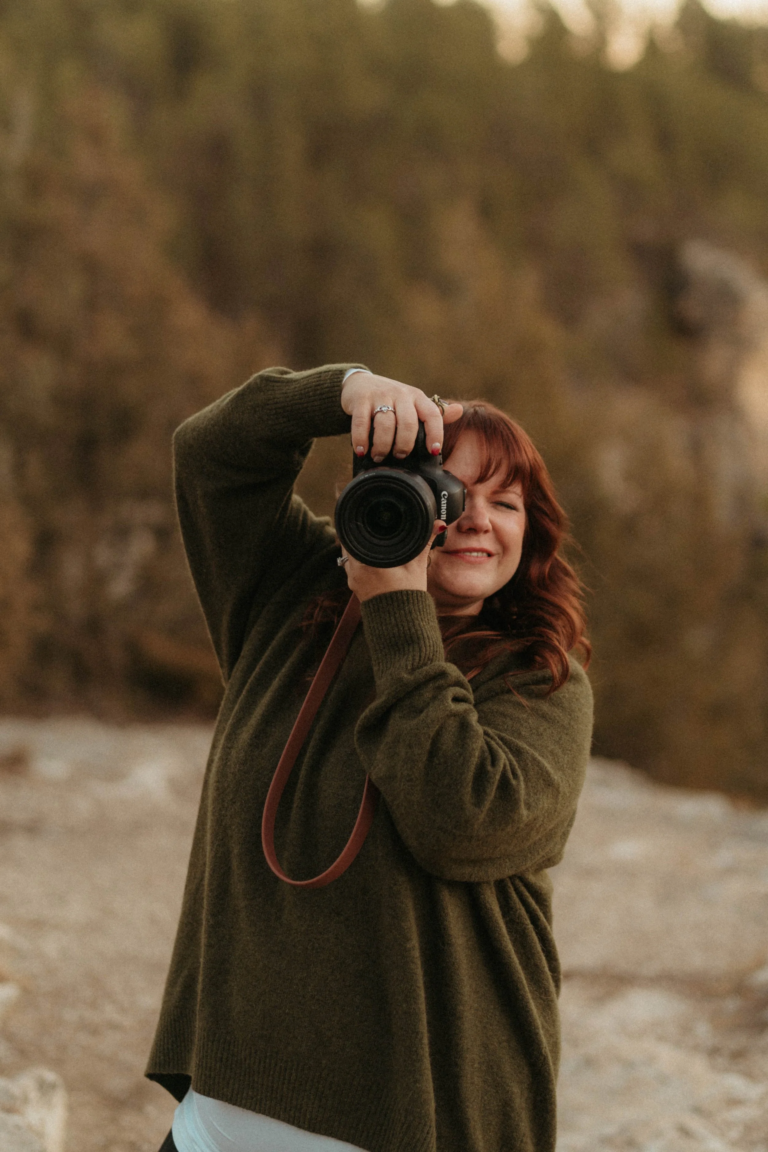 Woman with red hair taking a photo with a Canon camera outdoors, smiling, wearing a green sweater, with a blurred natural background.