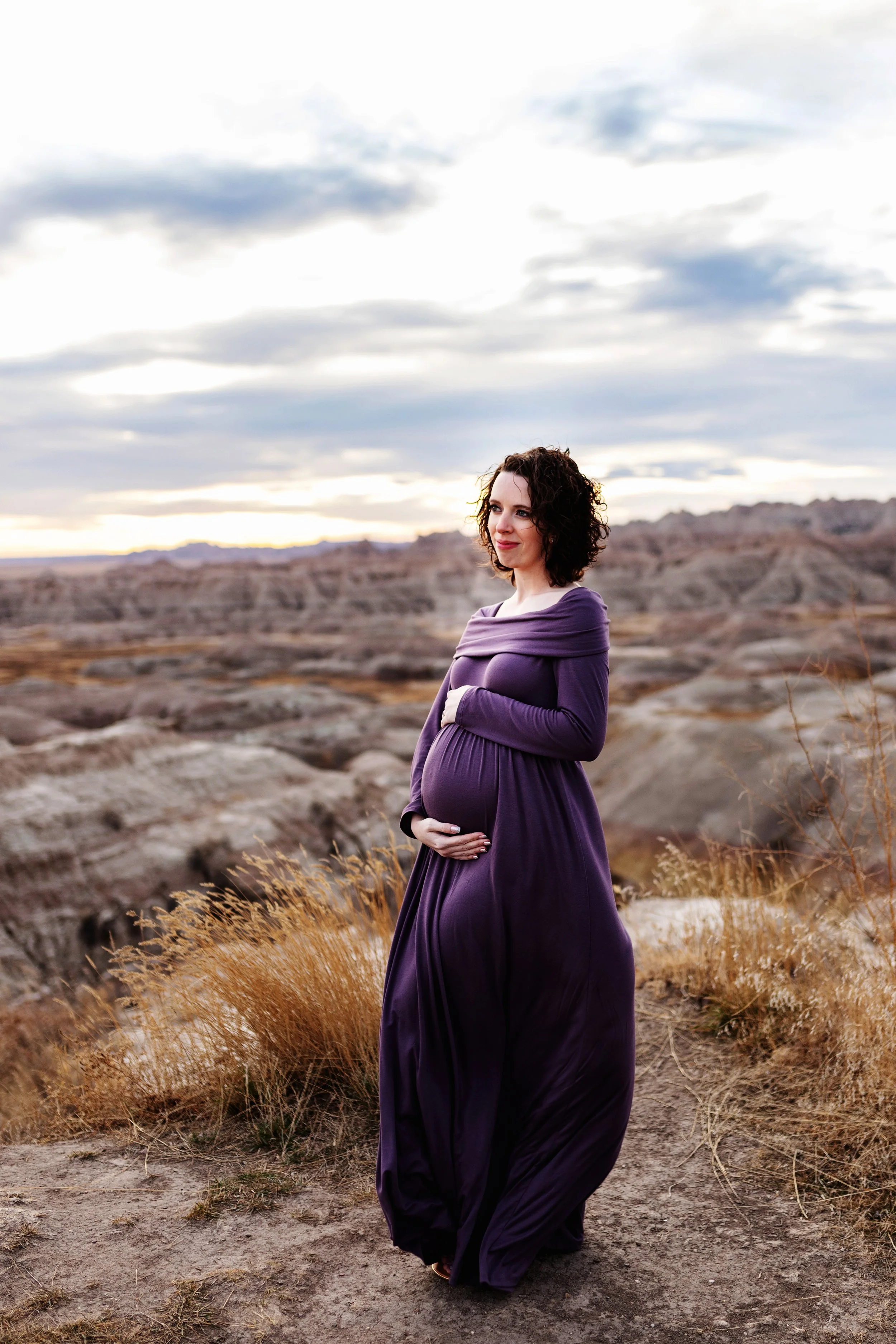 A pregnant woman in a long purple dress standing outdoors in a desert landscape with hills and shrubs, under a cloudy sky at sunset.