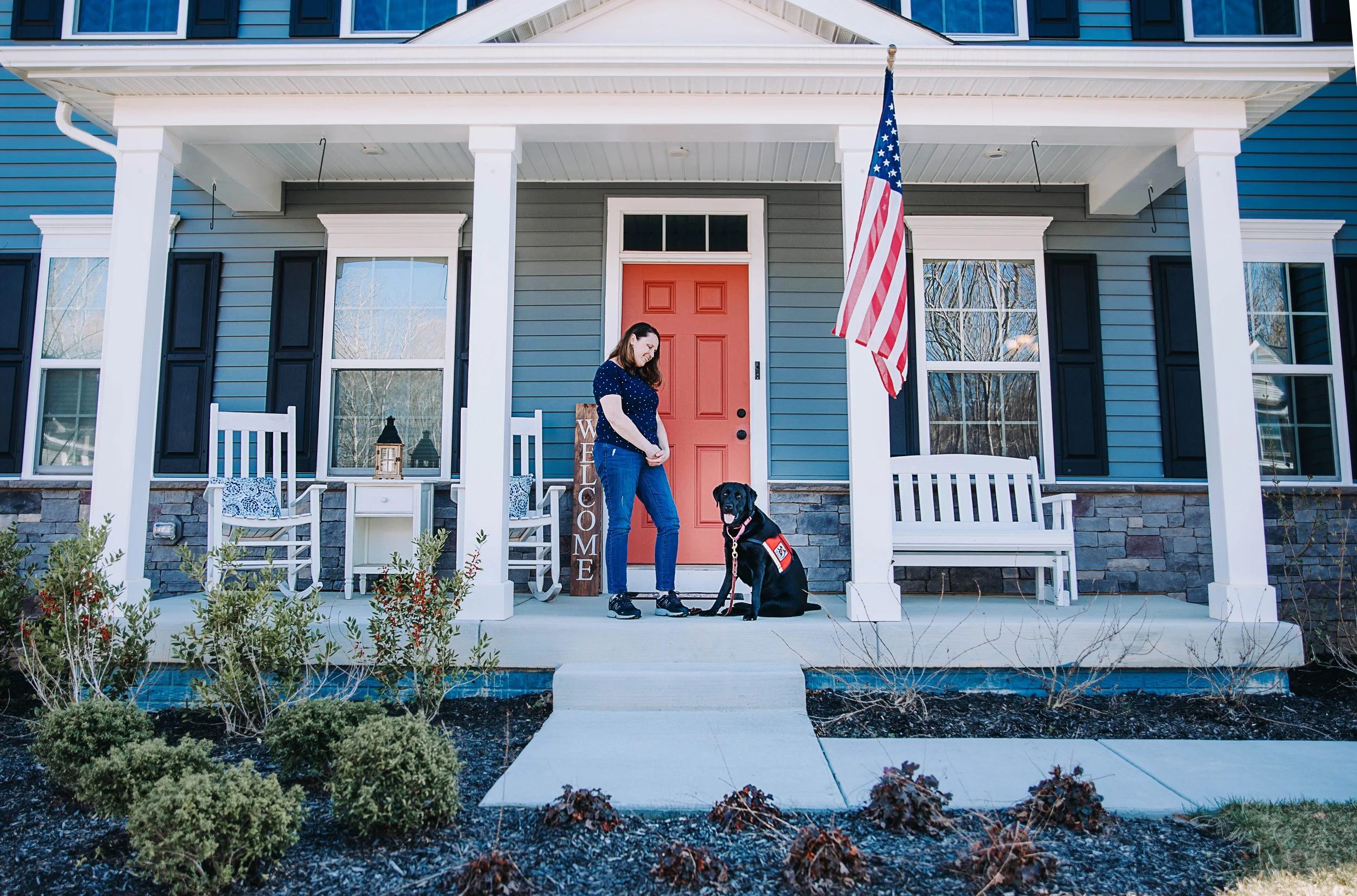 A woman with a service dog sitting at a house front porch. The house has a red front door, blue siding, black shutters, and a porch with white benches and chairs. An American flag hangs beside the door, and a welcome sign stands nearby. The yard has 