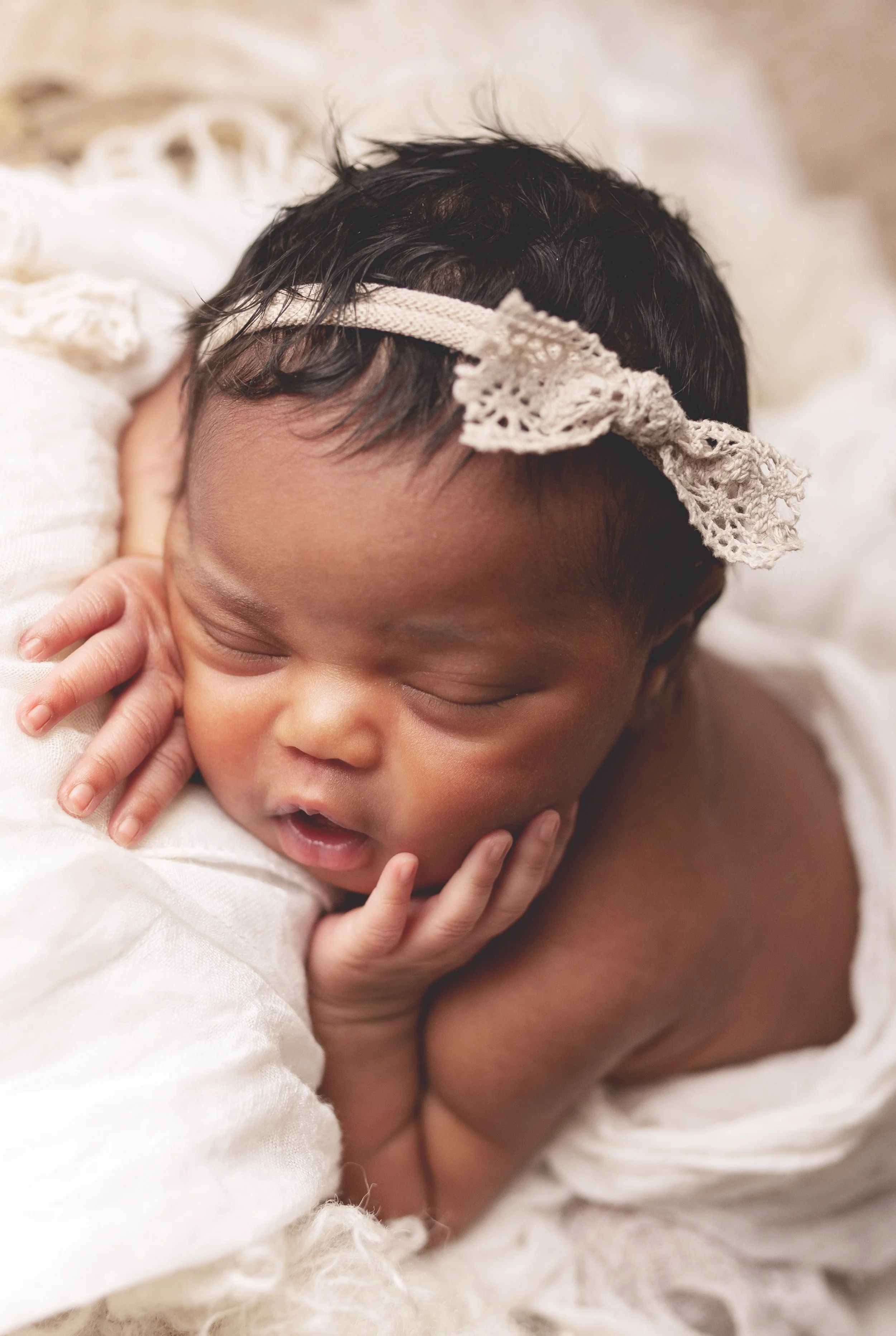 Close-up of a sleeping baby with dark hair, wearing a lace headband, resting on a soft, cream-colored blanket.