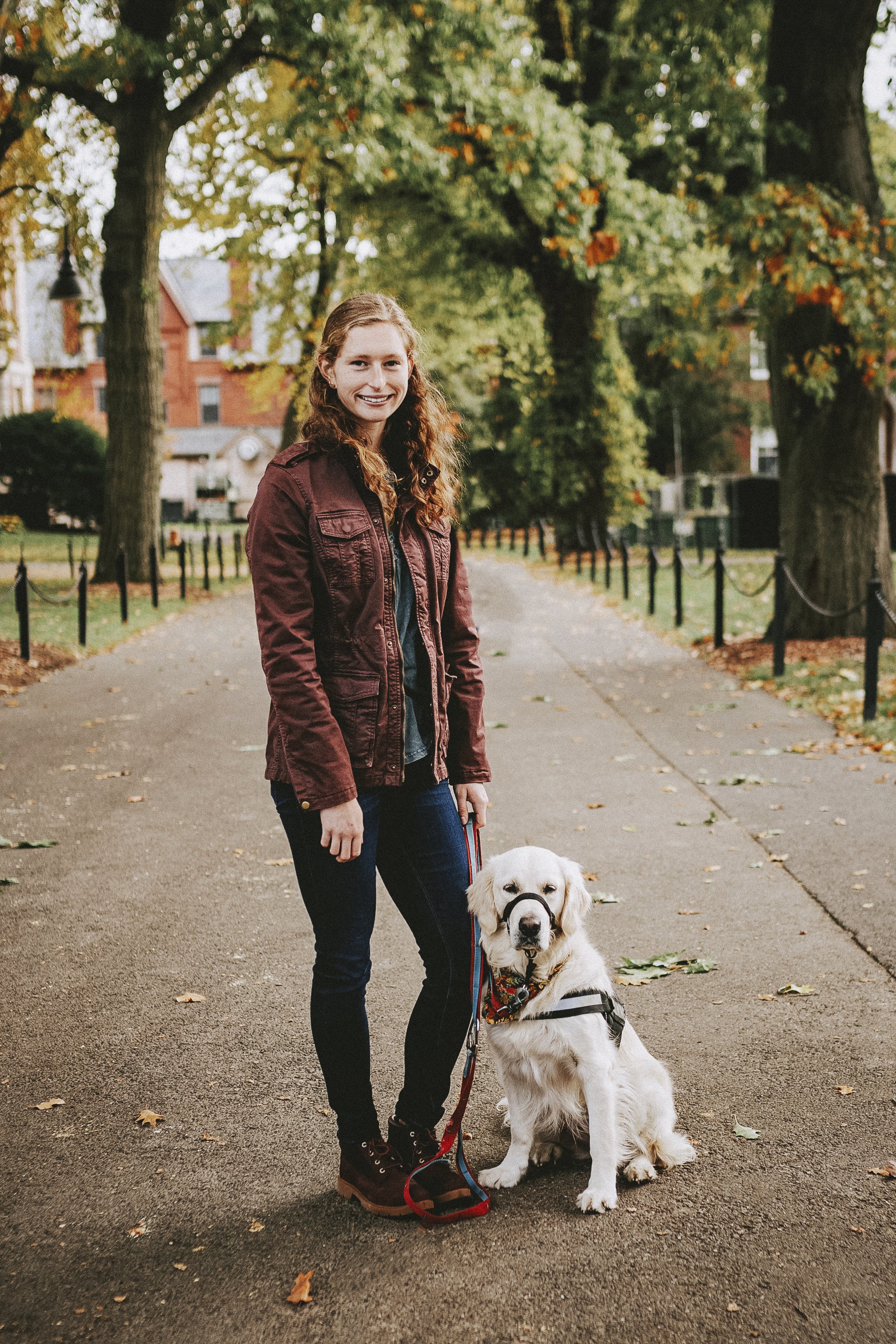 A young woman with curly red hair in a brown jacket, standing on a park path with a golden retriever wearing a harness and a bandanna, surrounded by trees with autumn foliage.
