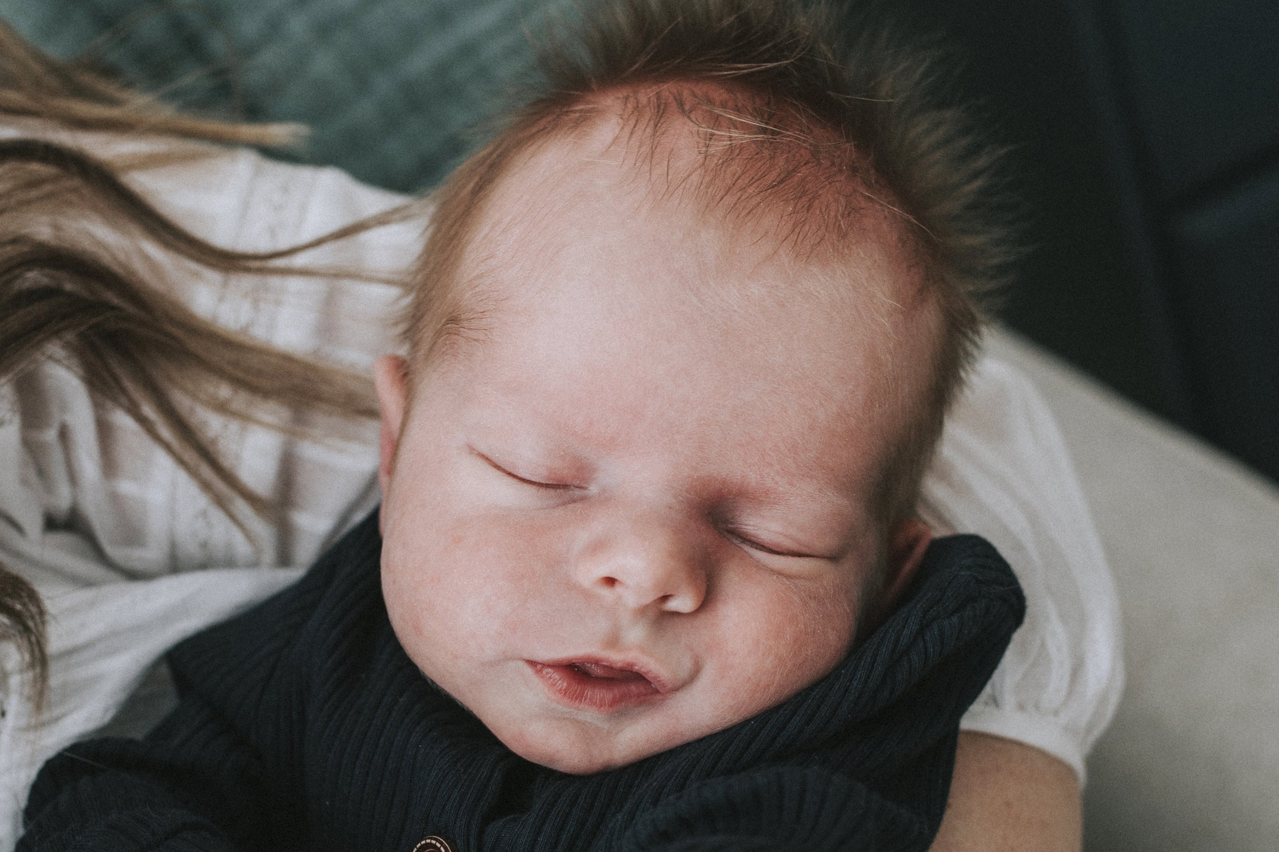 Close-up of a sleeping baby with reddish hair, wearing a dark-colored outfit, resting on an adult's arm.