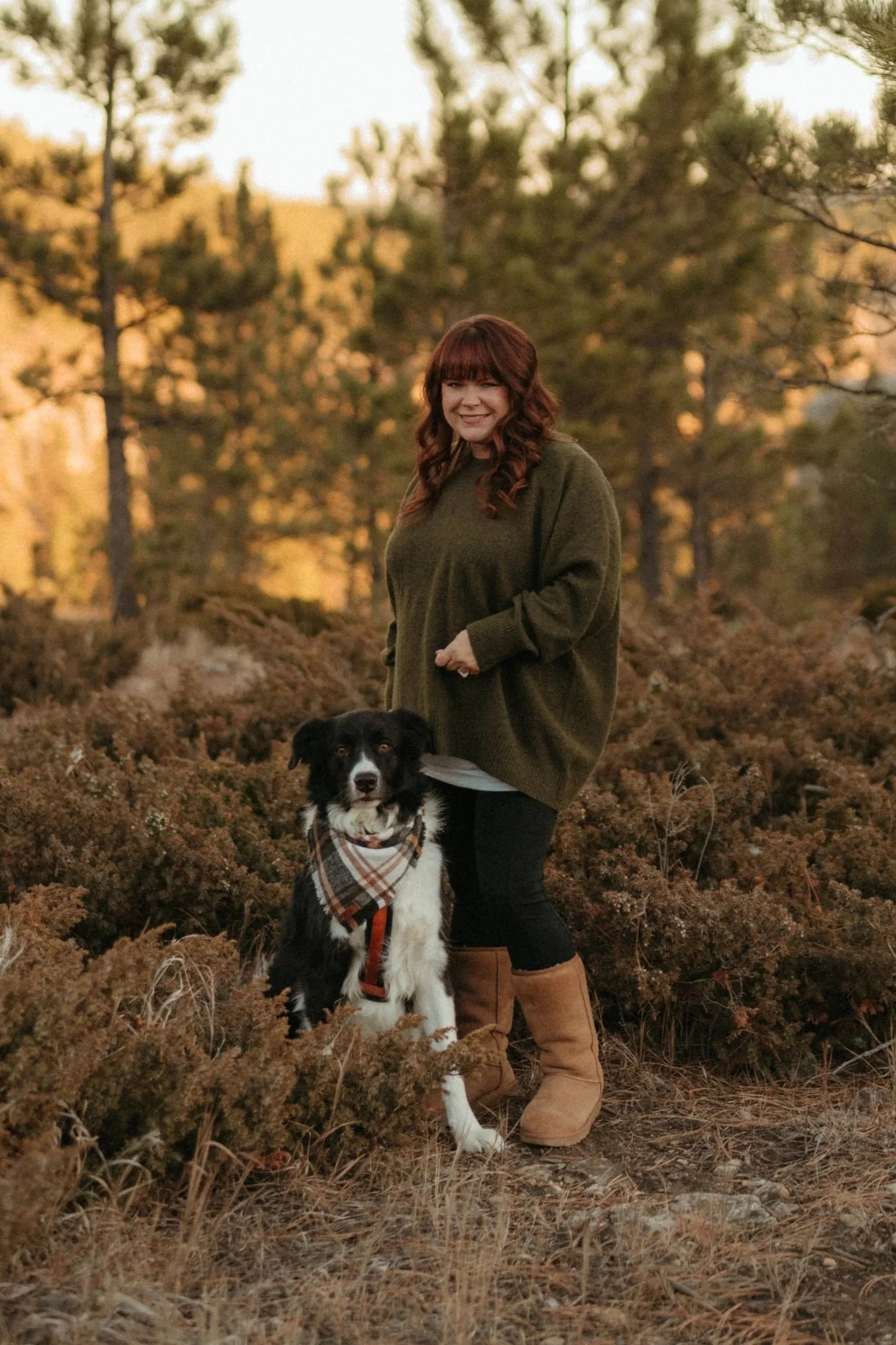 A woman with red hair in a green sweater standing outdoors on a dirt path with a black and white dog wearing a plaid bandana. The background features trees and a natural landscape during sunset.