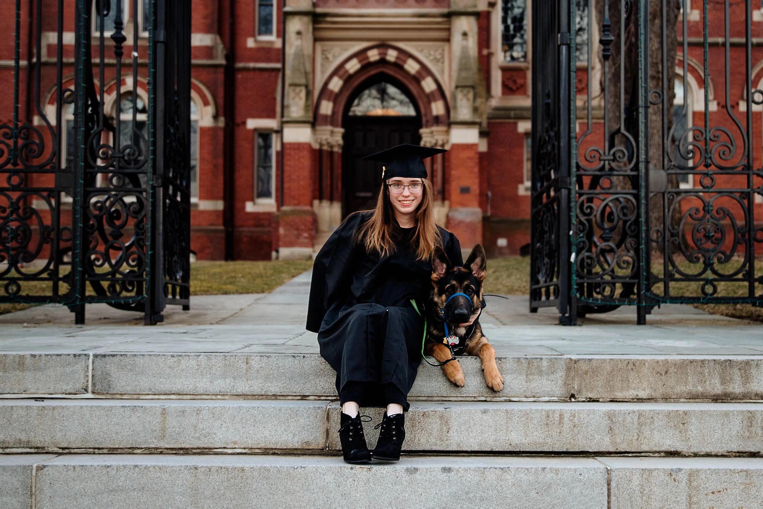 A young woman in a black graduation gown and cap sitting on steps with her German Shepherd dog in front of an ornate brick building with a gated entrance.